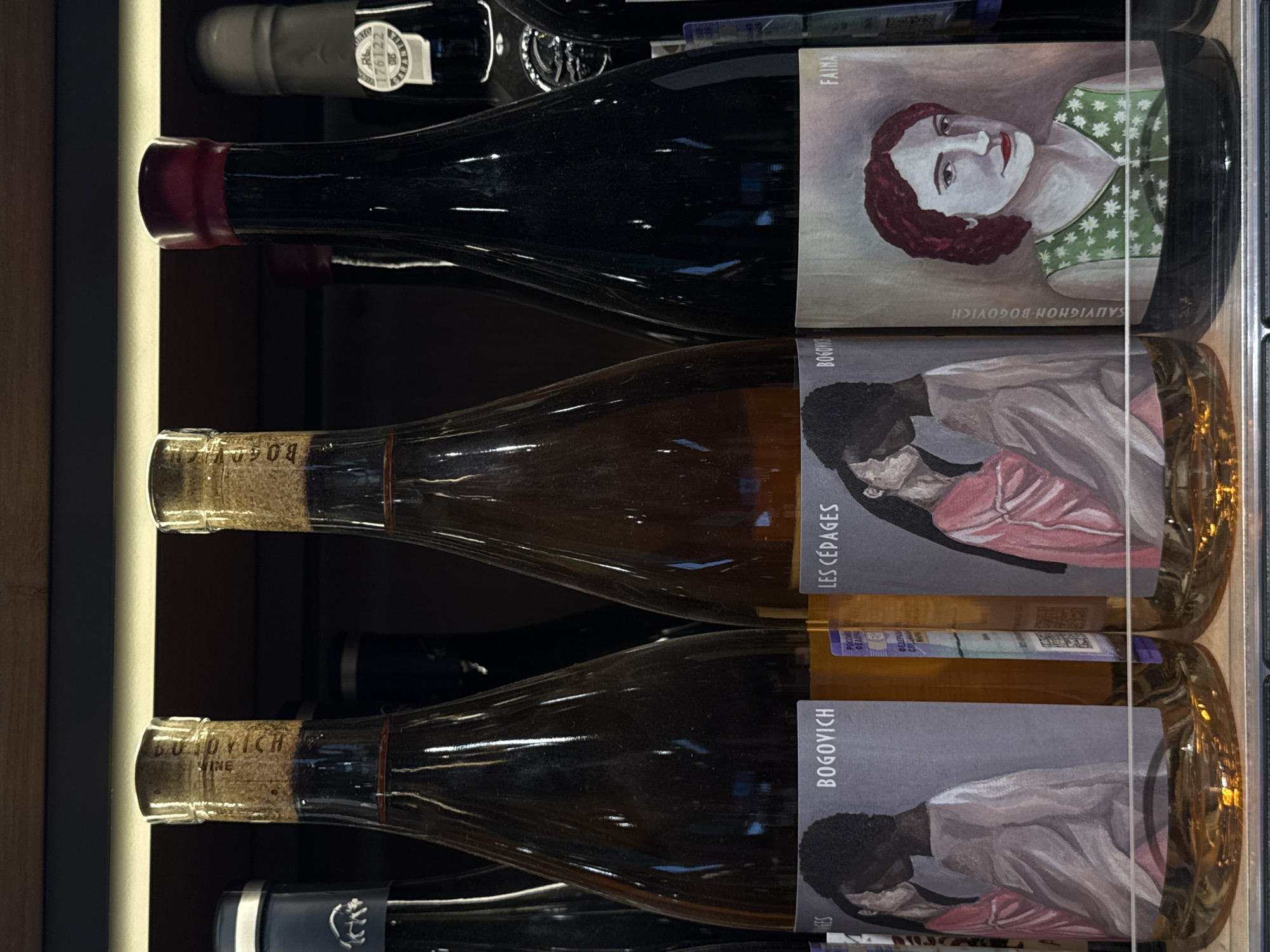 This is a close-up, eye-level shot of a shelf displaying bottles of wine in a Moscow, Russia establishment. The bottles are arranged in a row, with the focus on the labels of three bottles in the foreground. The leftmost bottle has a cork stopper and a label featuring a stylized illustration of two figures embracing, with the text "BOGOVYICH" prominently displayed. To its right, another bottle, also with a cork stopper, has a label that reads "LES CEPAGES" and shows a similar illustration to the first bottle. The third bottle, to the right, has a dark red wax seal and a label that says "FAINA" with a portrait of a woman. The background is dark and out of focus, suggesting an indoor retail or storage environment.