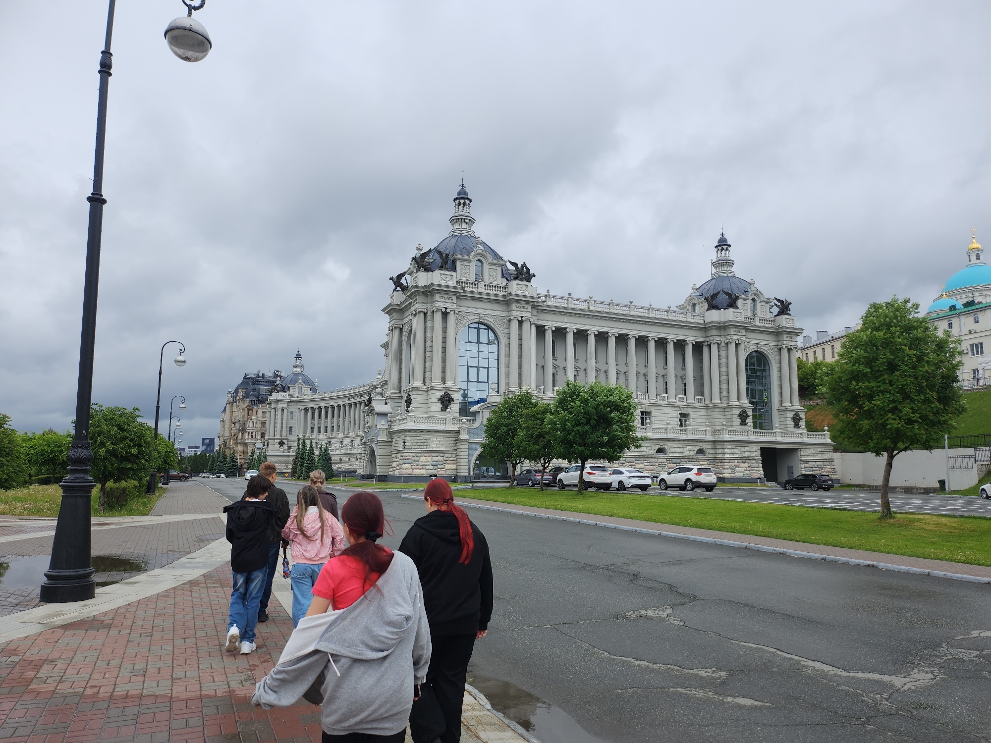 A group of six people is walking away from the viewer on a brick-paved sidewalk that transitions to asphalt, next to a road that is wet with recent rain. The location appears to be a city park, with a large, ornate building in the background. The building is light-colored, possibly white or grey, and has a prominent central section with columns and a large glass window. The building has domes with statues on top. A smaller building with a bright blue dome is visible to the right. Several cars are parked alongside the road near the large building. The sky is overcast and cloudy, suggesting a rainy day. The scene is in Unknown, Unknown.