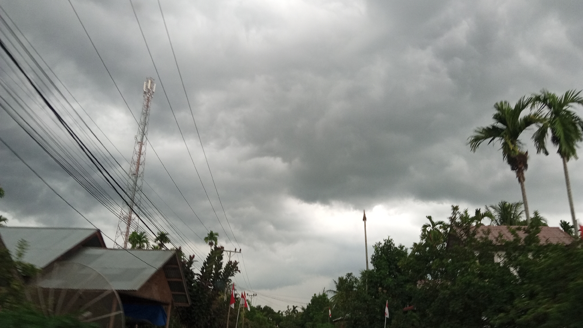 The image depicts a cloudy sky over a village, possibly in the city of Blang Mee, Indonesia. In the foreground, several houses with corrugated metal roofs are visible. A tall telecommunications tower stands prominently on the left side of the frame, intersected by numerous power lines. Palm trees are scattered throughout the scene, with two larger palm trees on the right. Several small flags, some appearing to be red and white, are planted near the houses and along the road. The overall atmosphere is overcast, suggesting impending rain or a storm. No people or animals are clearly visible in the image. The scene appears to be a rural or semi-rural residential area with infrastructure for communication and power.