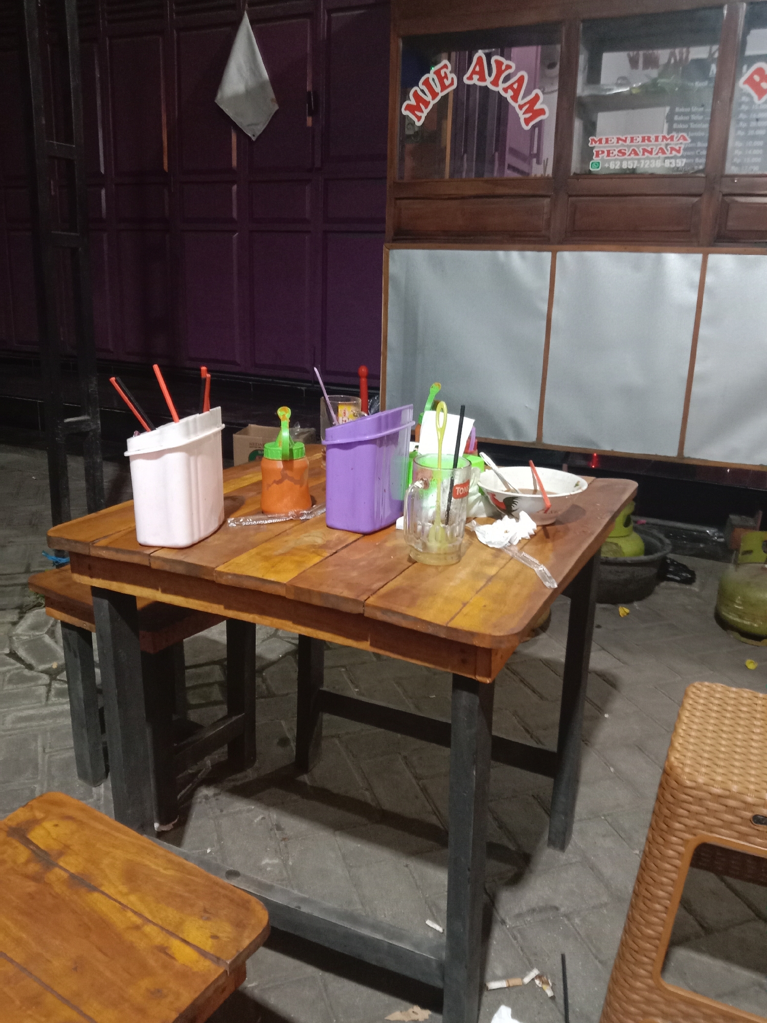 An outdoor eatery table and chairs are set up on a paved area in front of a building with a purple-paneled door and a wooden counter with glass windows. The counter has signs advertising "MIE AYAM" (chicken noodles) and taking orders. On the table, there are several items including a white container with orange and black utensils, a small orange sauce bottle with a green cap, a purple container, a clear glass with straws and a straw, a bowl with a spoon and fork, and a used napkin. Behind the table, to the right, are two propane gas tanks and a black basin. To the left of the table, a black metal table leg is visible, and a wooden bench is partially seen. A brown woven plastic stool is in the foreground to the right. The scene appears to be at dusk or night, as indicated by the lighting and shadows.