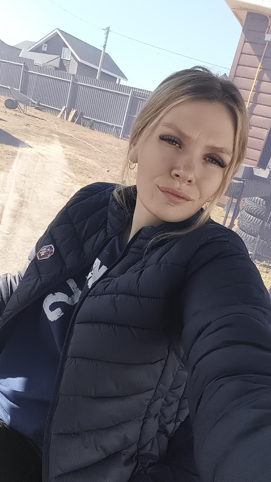 A young woman with light blonde hair, wearing a dark blue puffer jacket and a blue shirt with a white design, is taking a selfie. She is facing the camera with a slight smirk. In the background, there is a fenced-off dirt area with a house and other buildings visible. There are some stacked tires behind her to her right. The location appears to be in a rural area. Without additional information, pinpointing the exact location as Русский Вожой, Russia, is not possible.
