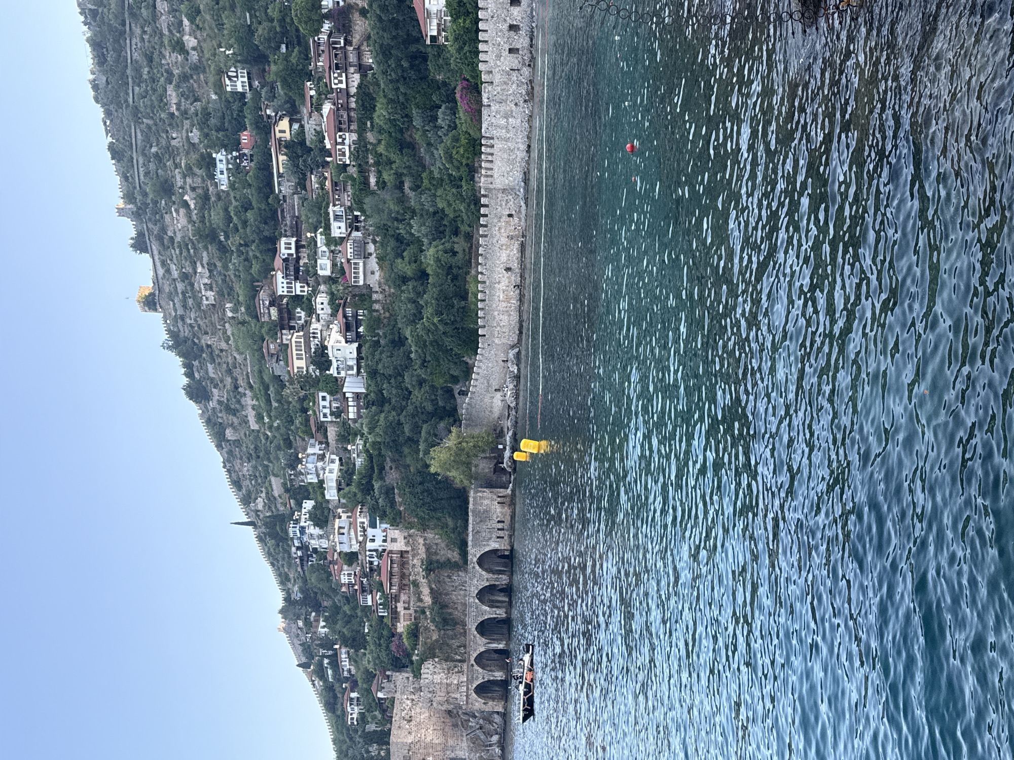 This is a vertical, low-angle, eye-level shot of a coastal town built on a steep hillside overlooking a body of water. The town features numerous white buildings with red-tiled roofs, nestled among lush green trees that cover the hillside. A stone wall, likely a historical fortification, runs along the edge of the town, adjacent to the water. The water is a deep blue-green, with ripples and whitecaps indicating a gentle breeze. A few yellow buoys are visible in the water near the wall. In the lower right portion of the frame, a small boat with two people is visible, navigating the water. The sky is a clear, light blue, suggesting it is daytime with fair weather. The overall scene evokes a sense of tranquility and picturesque coastal living. The architectural style of the buildings and the presence of a historic stone wall suggest a location with a rich history, consistent with the context of Alanya, Turkey. There is no visible text in the image.