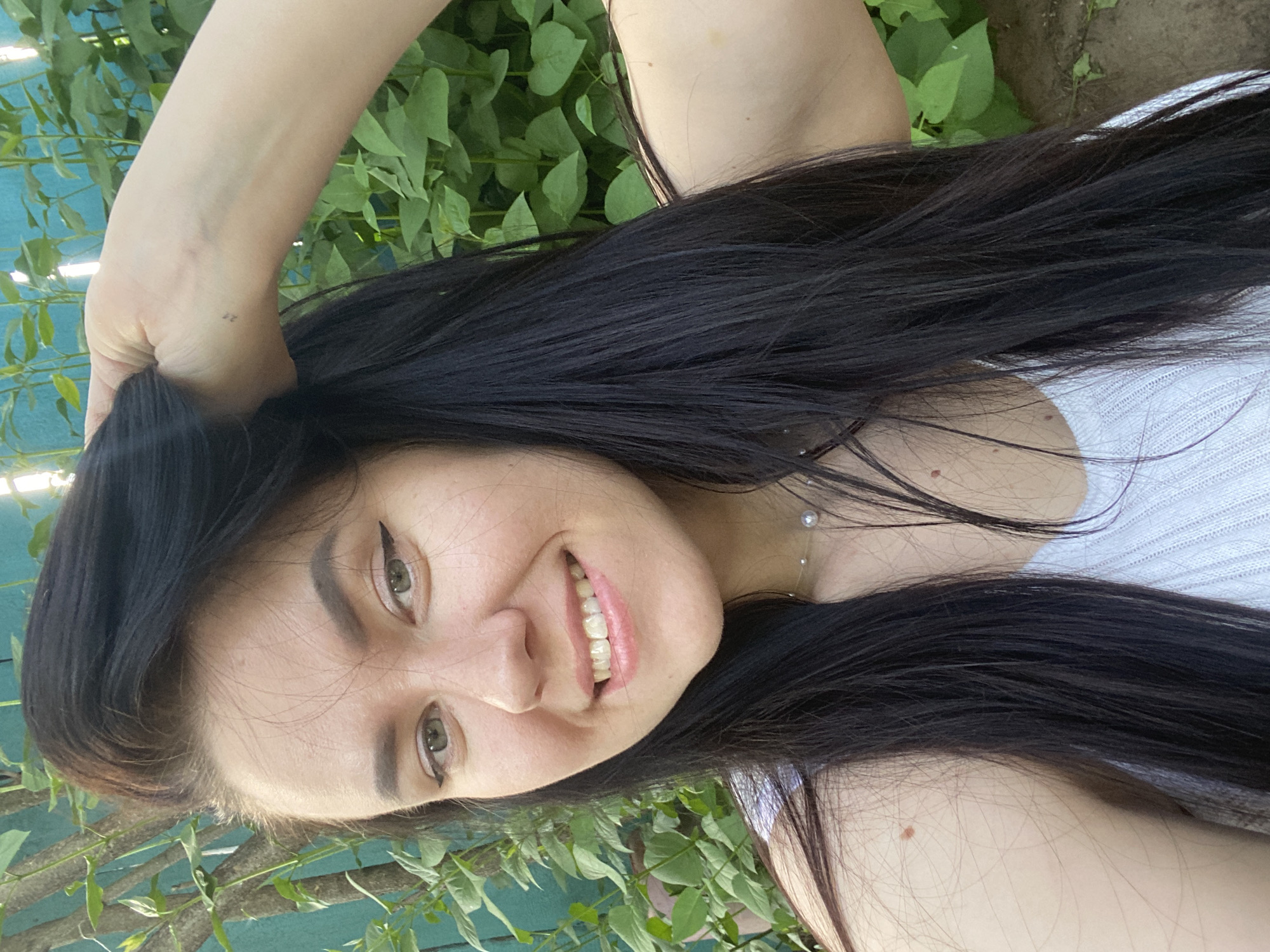 A young adult female is depicted outdoors in a close-up, waist-up selfie format, presumed to be in the city of Chaikovsky, Russia. She has fair skin, long, straight, dark hair, and light green eyes with dark winged eyeliner. Her eyebrows are dark and well-defined. She is smiling, revealing her upper teeth. Her left hand is raised to her head, with fingers partially visible in her hair, near her left temple. A small, dark tattoo, appearing to be the numeral "2" or a similar symbol, is present on her left wrist. She is wearing a white, ribbed knit sleeveless top and a delicate necklace featuring two pearls on a transparent string. Multiple small moles are visible on her shoulders and chest. The background consists of dense green foliage, with segments of a turquoise or light blue wooden fence visible behind the vegetation. The scene is illuminated by natural, even daylight.