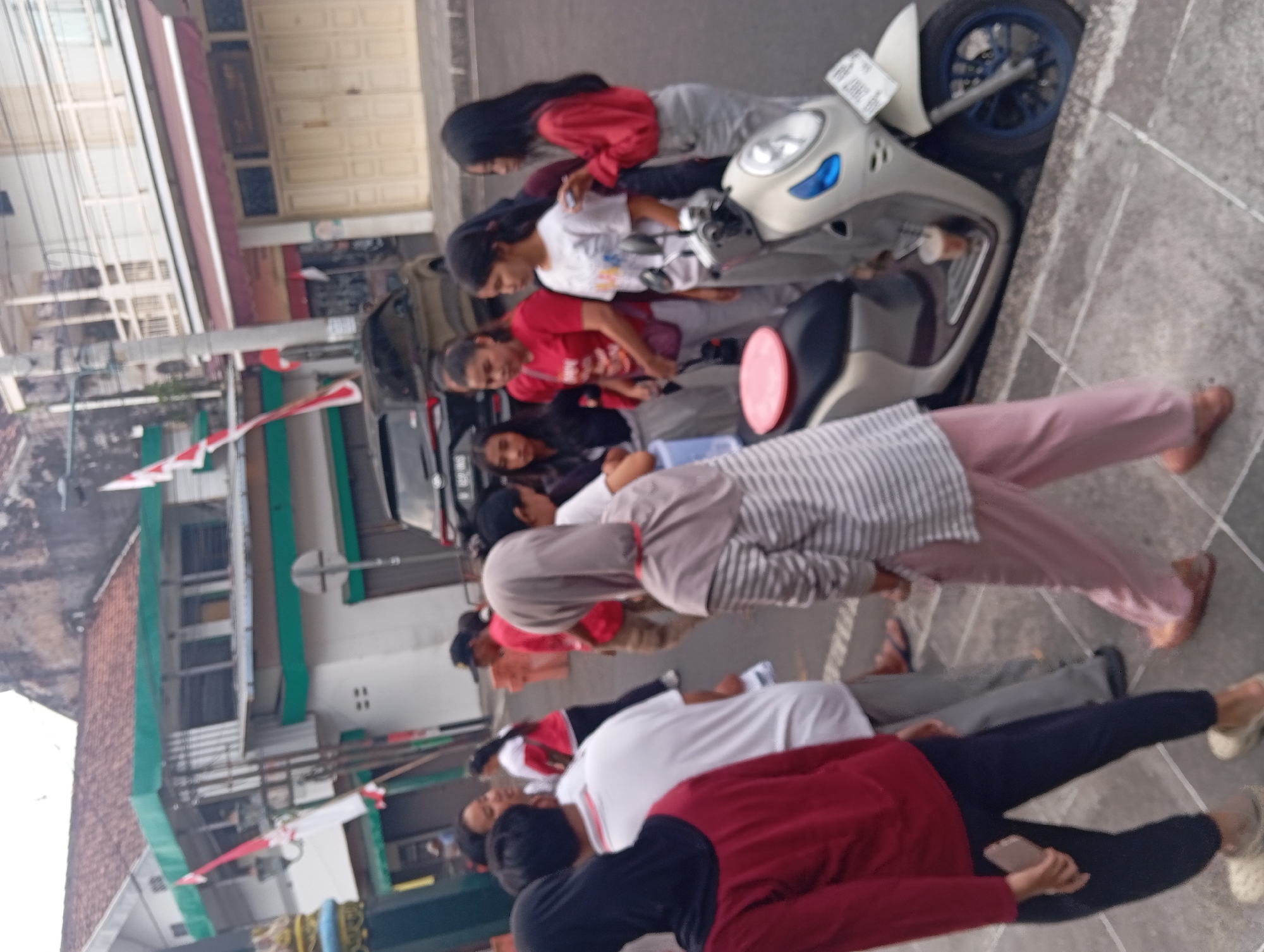 In Yogyakarta, Indonesia, a group of individuals stands near a street corner, with buildings and a parked motorcycle visible. A woman in a red shirt and grey pants stands next to a motorcycle, looking at her phone, while a group of younger women in red and white shirts also stand nearby. One woman in a grey hijab and striped shirt walks away from the camera. A man in a red shirt and beige pants stands to the right. The street is paved with stone, and buildings with red tiled roofs and white walls line the street. Indonesian flags are visible on poles. A black car is parked behind the group.