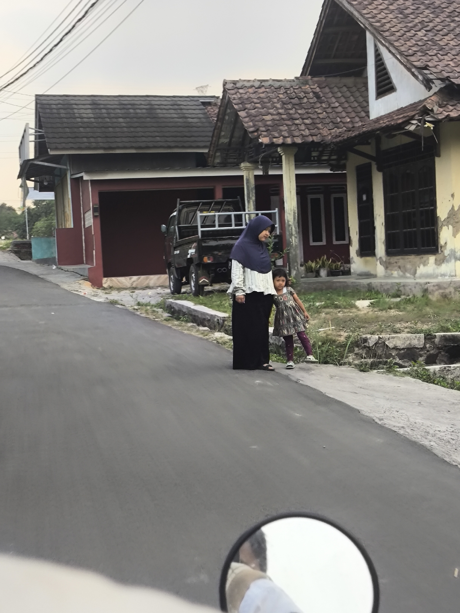 The image depicts a residential street scene in Cilegon, Indonesia, with two individuals, a woman and a child, standing on the side of the road. The woman, wearing a blue hijab and a long black skirt, stands next to a young girl dressed in a patterned dress and purple leggings. They are positioned on a paved sidewalk in front of a house with a yellow facade and a tiled roof. A dark-colored pickup truck is parked on the street nearby. The environment includes several buildings with tiled roofs, some appearing older and in disrepair, as well as vegetation and electrical wires overhead. The perspective suggests the photo was taken from a vehicle, possibly a motorcycle, given the presence of a rearview mirror in the foreground reflecting a person's head and shoulder.