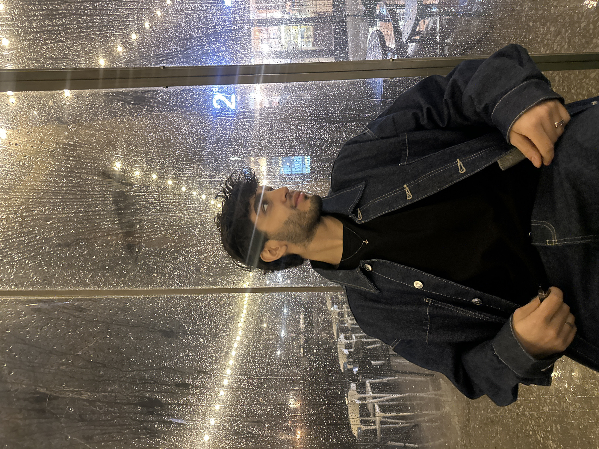 A medium shot of a man with dark hair and a beard, wearing a dark denim jacket over a black shirt, looking upwards and to the left. He's standing in what appears to be a bus stop or a shelter. Behind him, the transparent wall is covered in raindrops. Through the raindrops, blurred lights and a building with the letter "2" visible can be seen. The photograph is taken indoors, and the location is the city of Yerevan, Armenia.
