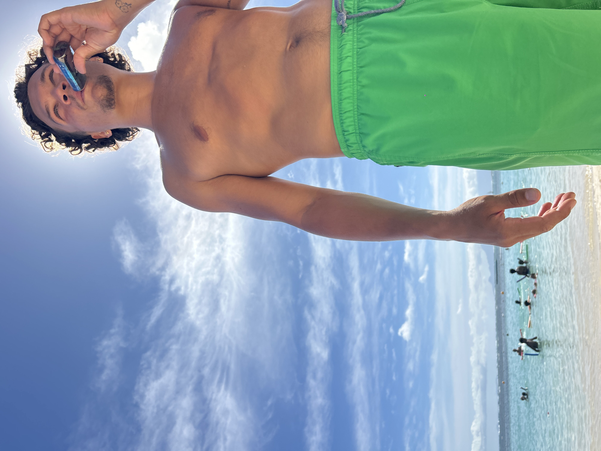 The image depicts a young man on a beach in Sainte-Anne, France, photographed from a low angle against a bright, sunny sky with scattered cirrus clouds. The man, shirtless, wears bright green swim trunks with a visible grey drawstring. He has dark, curly hair and a short goatee. He is positioned in the right half of the frame, looking upward, with the sun directly behind his head creating a strong backlight and a halo effect. He holds a cylindrical, dark blue and black electronic device, consistent with an e-cigarette or vape pen, to his mouth, appearing to inhale. Gold stud earrings are visible in both earlobes, a ring is on his left ring finger, and a faded cloud-like tattoo is on his left forearm. In the blurred mid-ground on the left, a calm, clear turquoise-blue body of water meets a light sandy shoreline. Approximately eight to ten indistinct figures are visible in the water, some appearing to be swimming or wading, with several using colorful flotation devices. A faint landmass is discernible on the distant horizon.