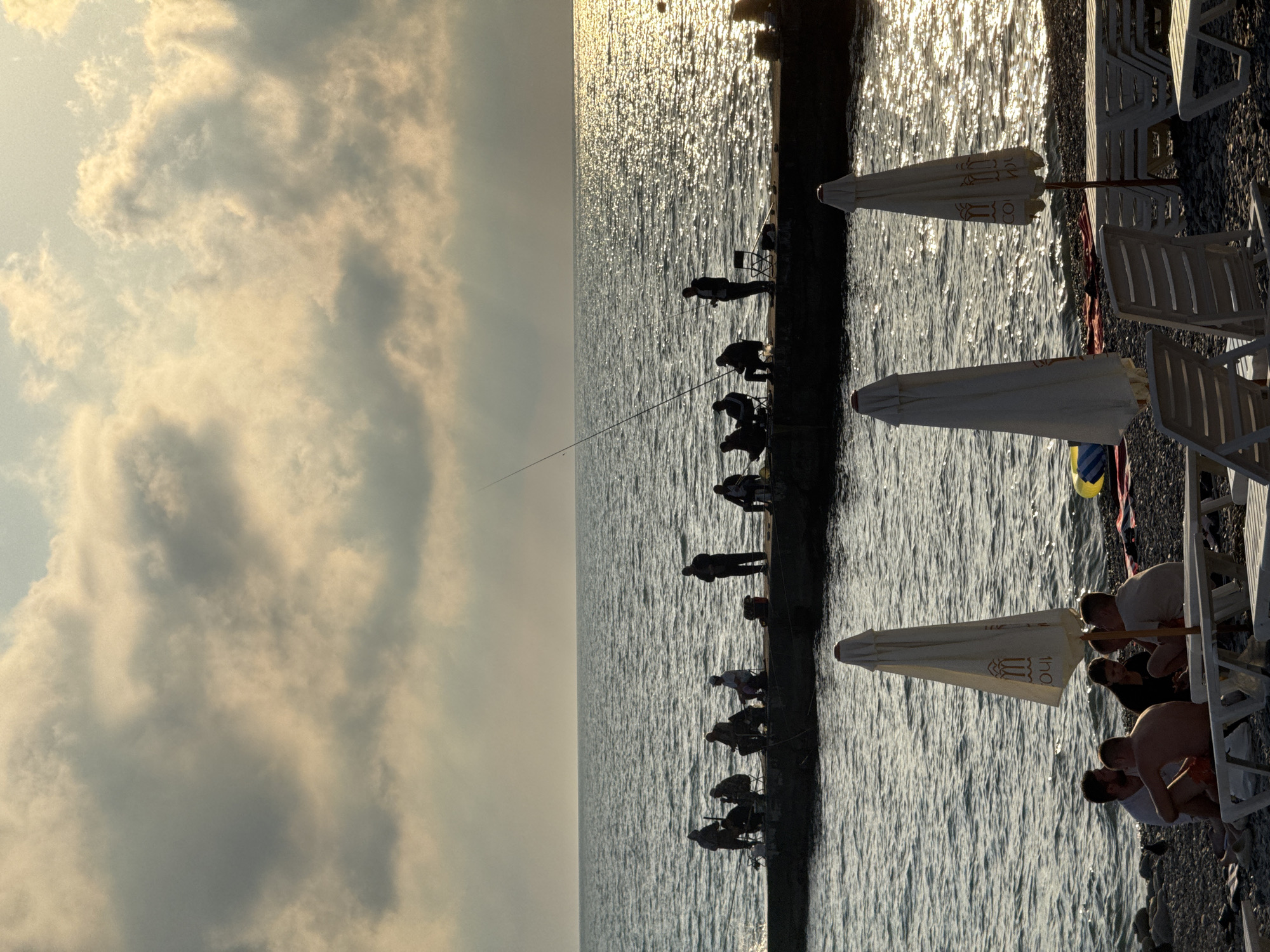 In Sochi, Russia, on a pebble beach under a cloudy sky, a group of people are fishing from a pier. The sun is setting, casting a golden shimmer across the water. In the foreground, three beach umbrellas are closed, and several lounge chairs are arranged on the beach. Some individuals are seated and appear to be conversing, while others are reclining on the chairs. The overall atmosphere is tranquil and relaxed, suggesting a peaceful evening by the sea.
