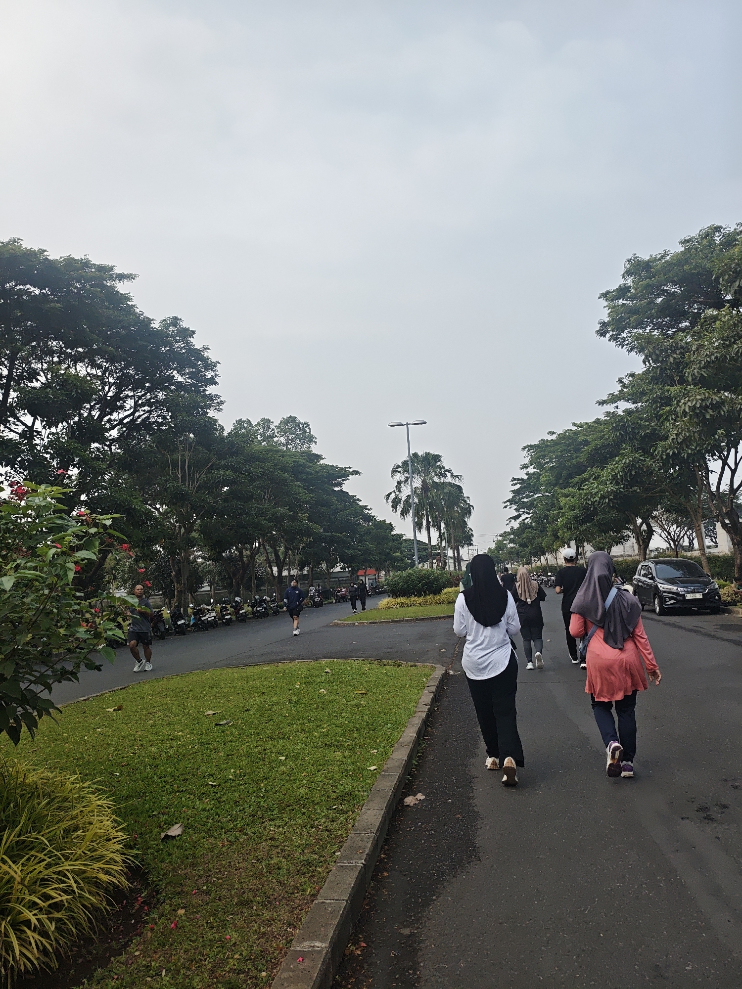 The image depicts an outdoor scene with a group of people walking or jogging on an asphalt road lined with trees. The setting appears to be a park or a residential area with well-maintained greenery and a row of parked motorcycles on the left side of the road. In the background, there are palm trees and a street lamp, suggesting a tropical or subtropical climate. The sky is overcast.
The foremost figures in the image are two women wearing headscarves and athletic attire, walking away from the camera on the right side of the road. The woman on the far right is wearing a coral-colored top and dark pants, while the woman to her left is wearing a white long-sleeved shirt and black pants. Behind them, further up the road, other individuals are also visible walking or running.
On the left side of the road, a man in athletic shorts and a shirt is jogging towards the camera. A grassy area with some vegetation is present between the road and the line of motorcycles. A dark-colored sedan is parked on the right side of the road in the distance. The overall impression is of a recreational activity taking place in a moderately urban environment.
Based on the information provided, this scene is located in the city of Bacin, Indonesia.