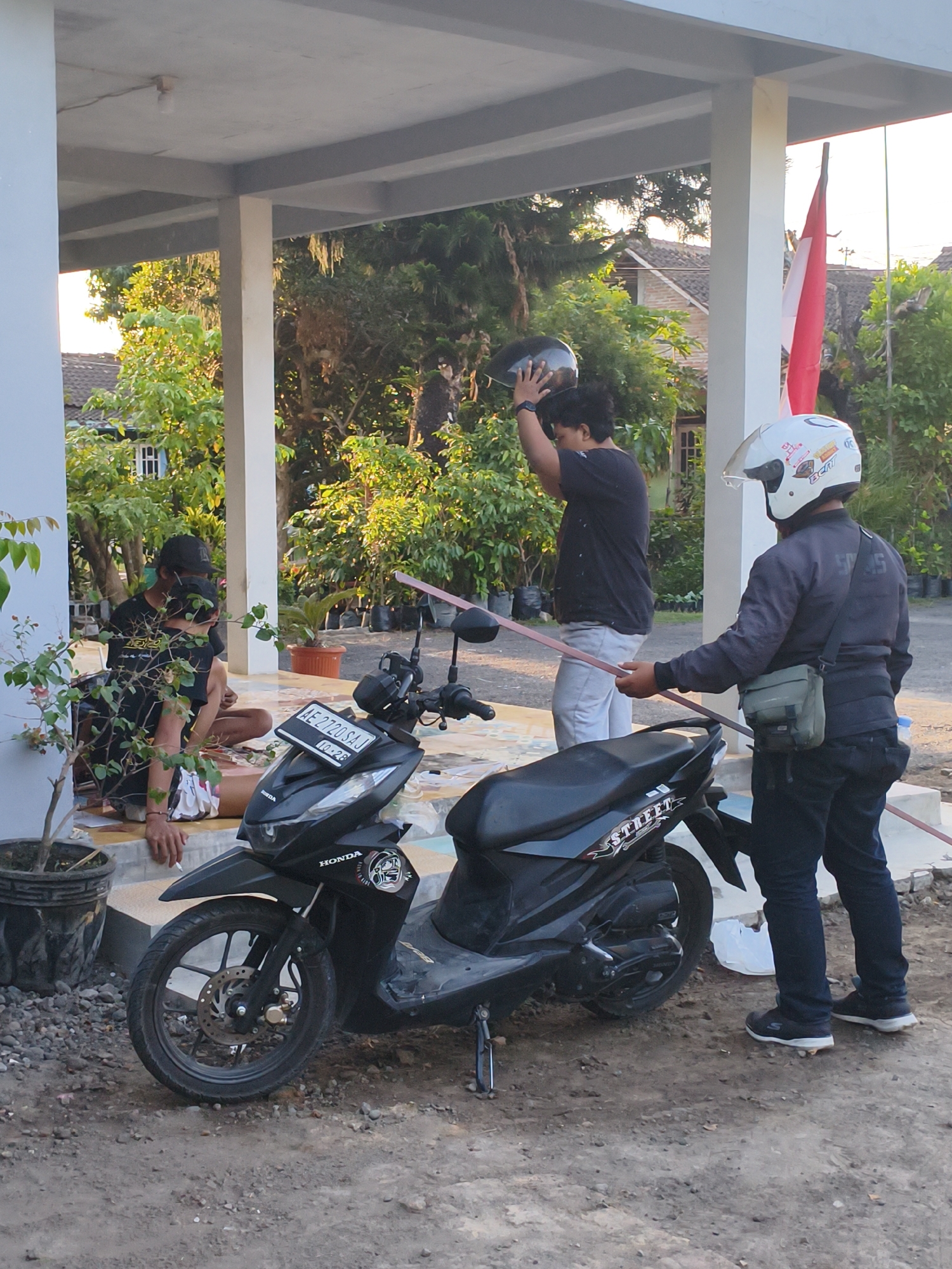 Two men and a motorcycle are in front of a house in Berbah, Indonesia. The man on the left is wearing a black t-shirt and a black baseball cap. He is seated with his legs crossed and is holding a cigarette. The man on the right is wearing a dark jacket, dark pants, and a white helmet. He is standing next to the motorcycle and holding a pink pole. The motorcycle is black and has the word "STREET" on the back. The background includes trees, houses, and a red and white flag. The sun is shining and casting shadows.