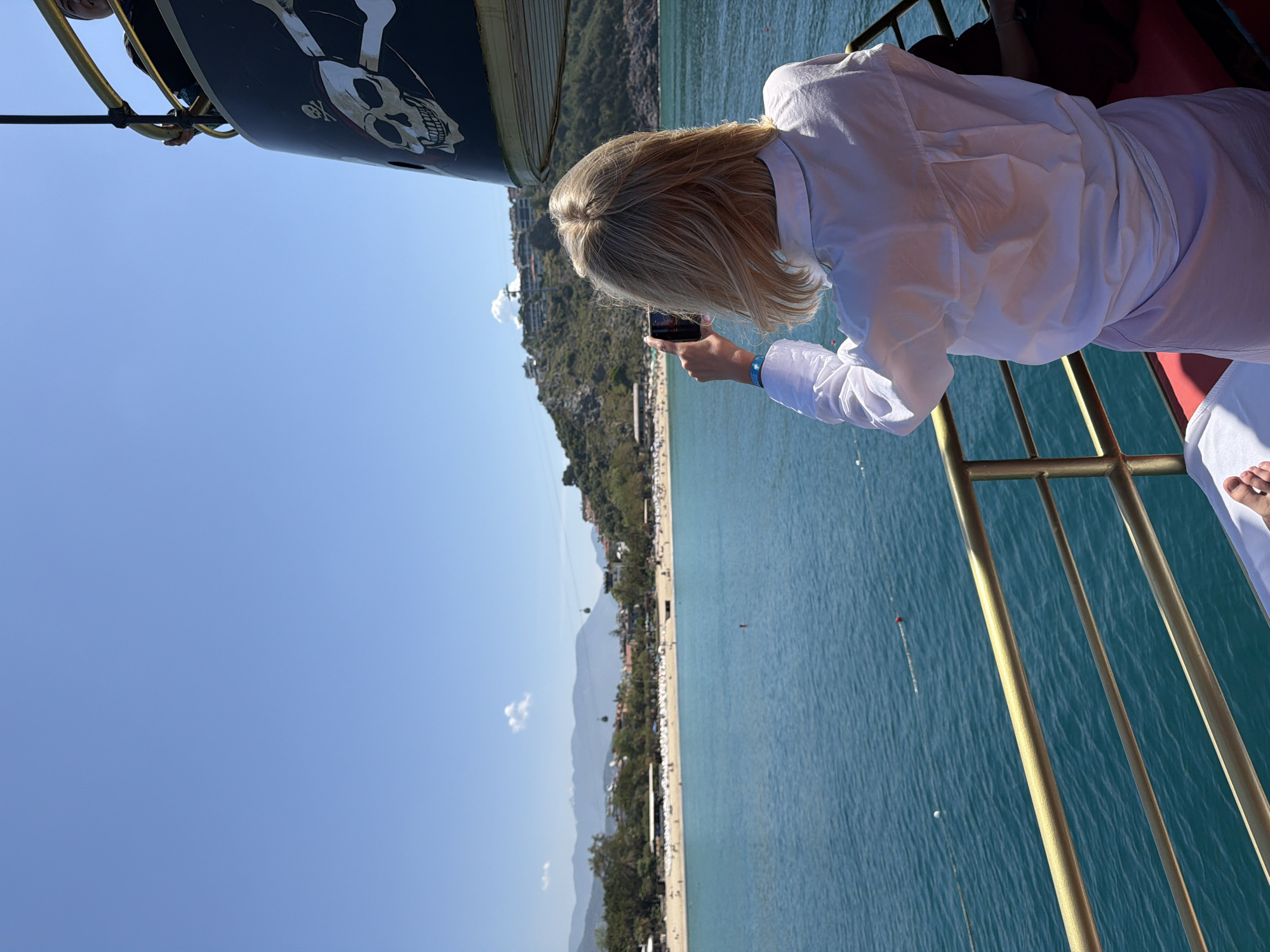 The image depicts a scene from what appears to be a boat trip in Alanya, Turkey. A fair-haired woman in a white shirt and white pants is taking a picture with her smartphone. The background includes a clear blue sky, a stretch of the turquoise Mediterranean Sea, and a long sandy beach lined with trees and buildings. In the distance, mountains can be seen. The boat is adorned with pirate-themed decor, including a skull and crossbones.