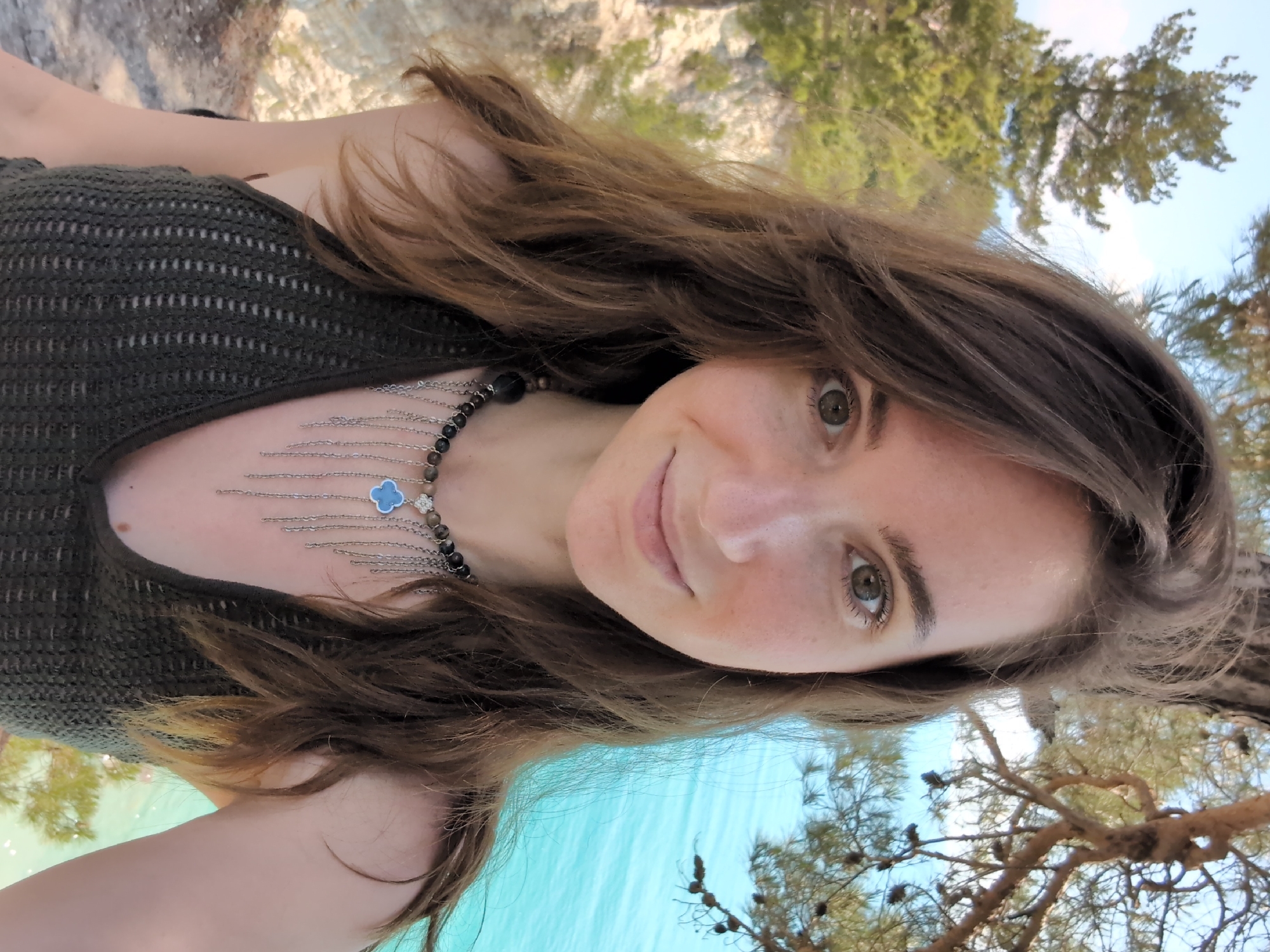A young woman with long brown hair and light skin poses for a selfie outdoors, with a body of turquoise water and a cliff face in the background. She is wearing a dark green knitted top and a beaded necklace with silver chains and a blue clover pendant. The scene appears to be located in Divnomorskoye, Russia, possibly near the coast, with trees and a clear sky visible. The woman's face is slightly turned to her left, and she is smiling.