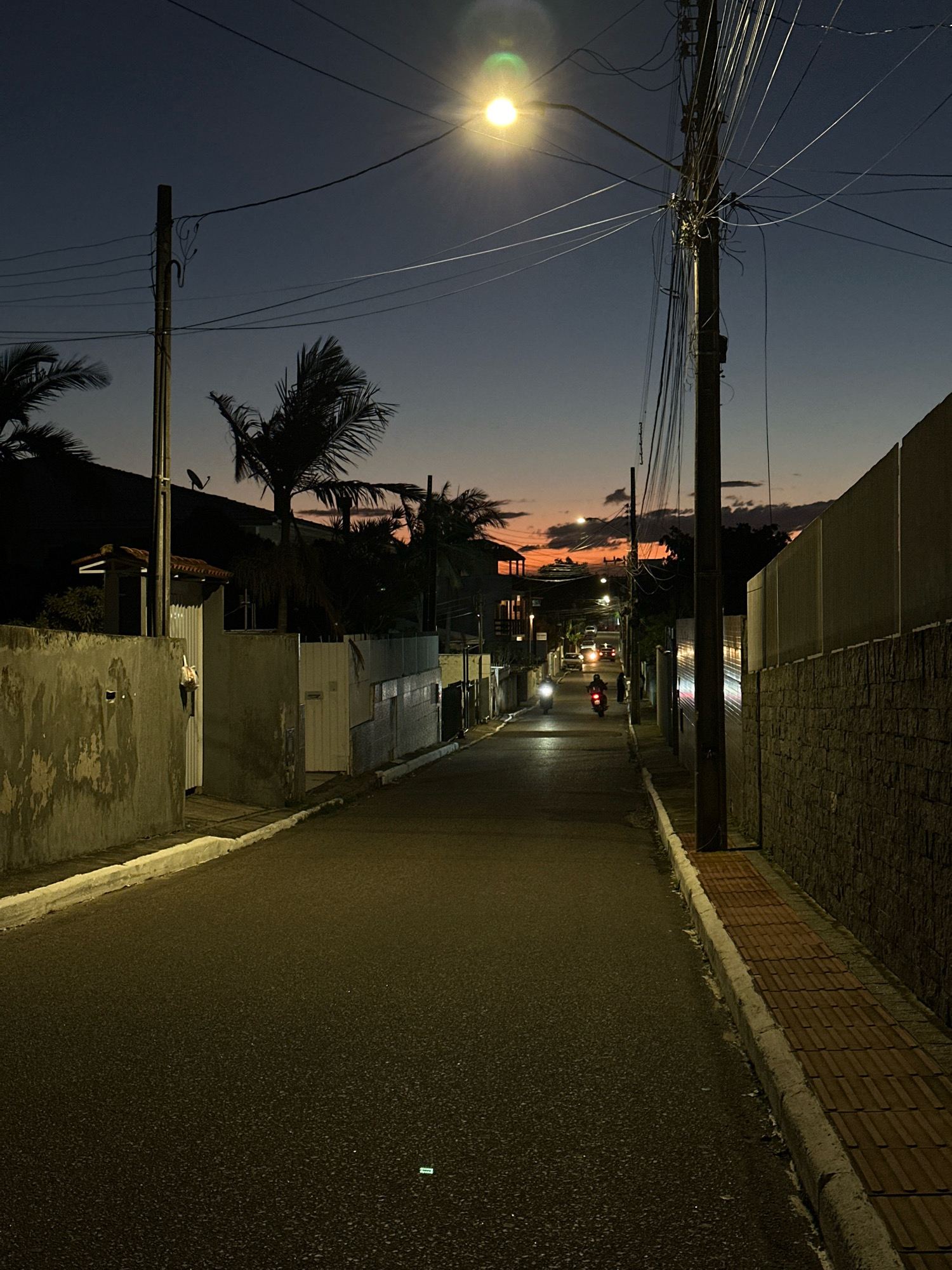 This image depicts a street scene in Florianópolis, Brazil, at dusk. The road is paved and curves slightly into the distance. On the left side of the road, there are concrete walls and fences of houses, with some vegetation, including palm trees, visible behind them. On the right, a wall constructed of dark bricks lines the street, with a textured brick walkway running along its base. A utility pole with a dense network of wires stands on the right side of the road. A streetlamp illuminates the scene from above and to the left, casting a bright glow. In the distance, two motorcycles are seen traveling down the street, their headlights illuminating the road ahead. The sky is a gradient of dark blue to orange and pink, indicating the sun has recently set, with some clouds scattered across it. The overall impression is one of a quiet residential street in a coastal Brazilian city as night begins to fall.