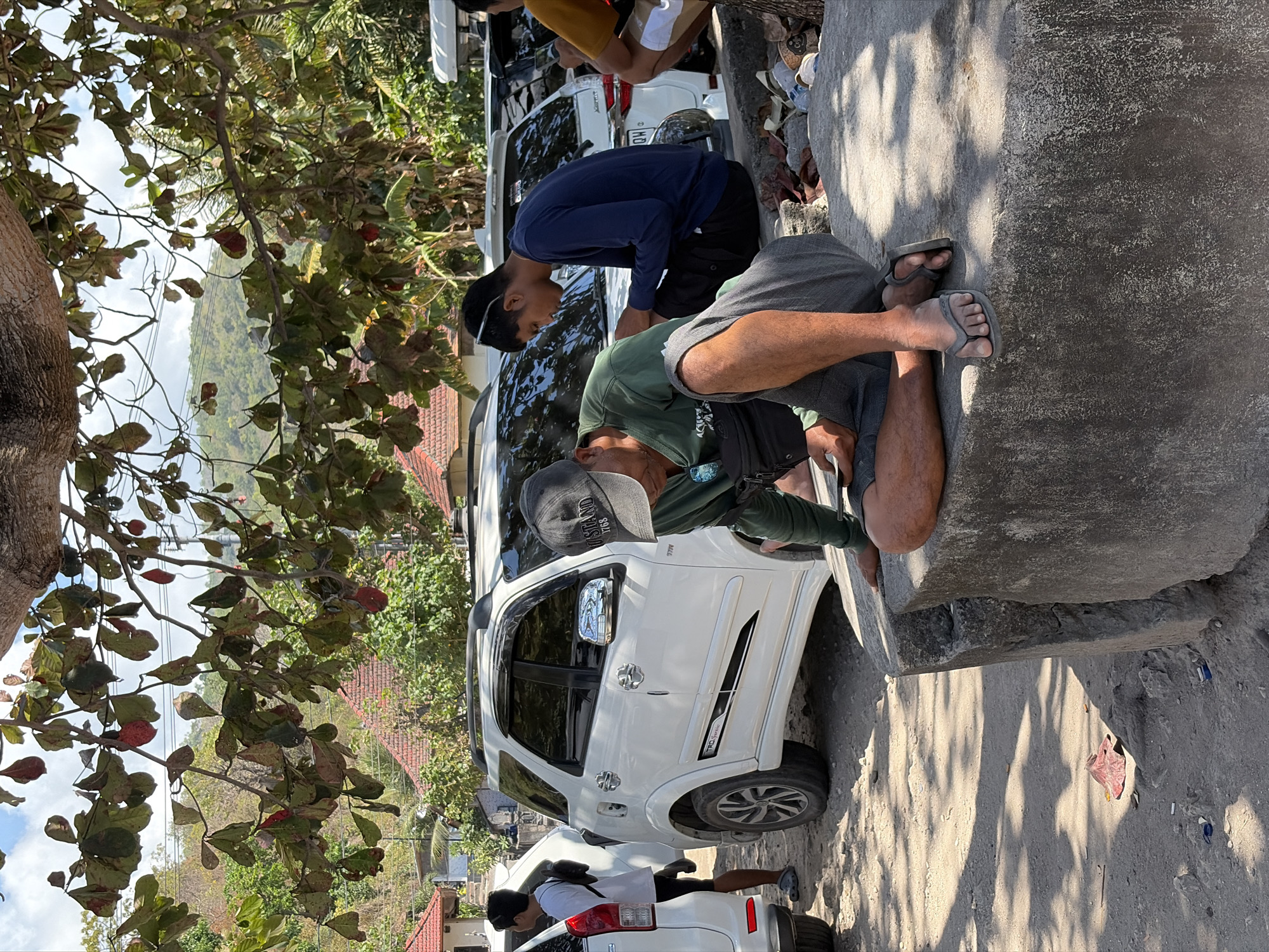 The image captures a street scene in Nusa Penida, Indonesia. In the foreground, a man wearing a green long-sleeved shirt, grey shorts, and flip-flops is crouched on a large concrete block, looking down at his phone. To his right, another man in a navy blue long-sleeved shirt and black shorts is also seated, looking at his phone. Behind them, two white SUVs are parked. A young man in a white t-shirt and black shorts stands near the left SUV. The background reveals tropical vegetation, houses with red tile roofs, and a hazy, mountainous landscape under a bright sky. The ground is dirt and gravel, with some scattered leaves and debris. The overall atmosphere is casual and relaxed, suggesting a warm, sunny day.