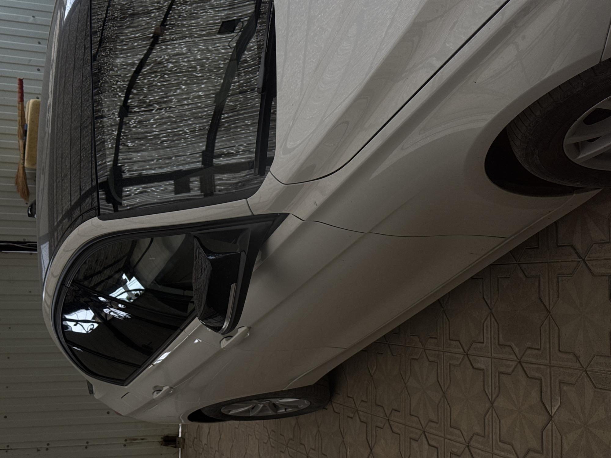A white sedan is parked indoors, likely in a garage or covered parking area. The car is angled with its side facing the viewer. The vehicle has tinted windows and a black side mirror. A broom with a yellow sponge is visible leaning against a corrugated metal wall to the left of the car. The floor is tiled with a geometric pattern. Reflections on the car's surface suggest an overhead structure, possibly a roof or awning. The scene appears to be lit by artificial light, as there are no strong shadows indicative of direct sunlight. There is no visible text in the image.
