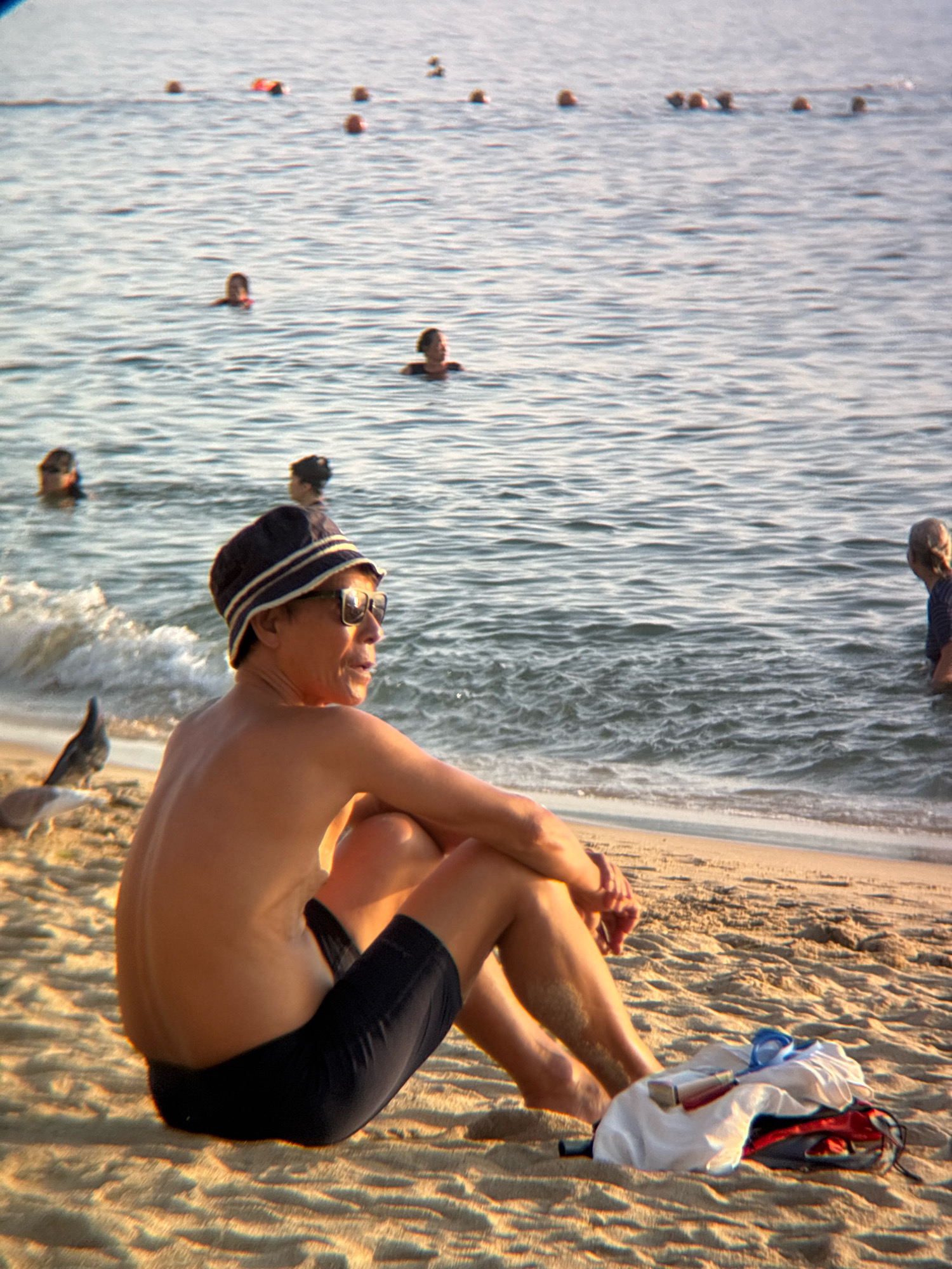 The image depicts a sunny day at a beach, likely in Nha Trang, Vietnam, given the presence of people swimming in the ocean and the overall tropical atmosphere. In the foreground, a person is sitting on the sand, facing away from the camera and looking out at the water. They are wearing a dark, striped bucket hat and sunglasses, and their body is bare from the waist up. They appear to be wearing dark swimming shorts. To the left of this person, a bird is perched on the sand. In the middle ground, several other individuals are visible in the shallow water, some with their heads above the surface. The water itself is calm with small waves lapping at the shore. In the background, the ocean stretches to the horizon under a clear sky. Scattered personal items, including a red and black bag and white clothing, are on the sand near the foreground subject.