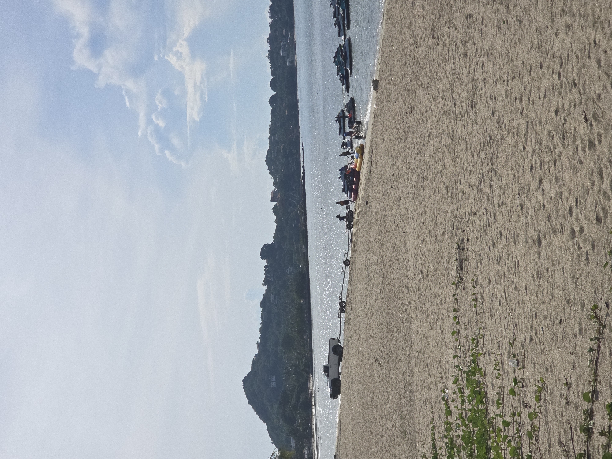 A bright, sunny day at a sandy beach in Baan Lamai, Thailand. The foreground is dominated by the beige sand of the beach, with some low-lying green vegetation growing in the lower left corner. In the midground, the calm, sparkling blue ocean meets the shore. A white pickup truck is parked on the sand near the water's edge, attached to a trailer with multiple axles. Several people are gathered around the truck and trailer, and further along the beach, a group of people are interacting with jet skis and other watercraft in the shallow water. In the background, a lush green, tree-covered hill rises from the coastline, with scattered buildings visible on its slopes and crest. The sky above is a light blue with scattered, fluffy white clouds.
