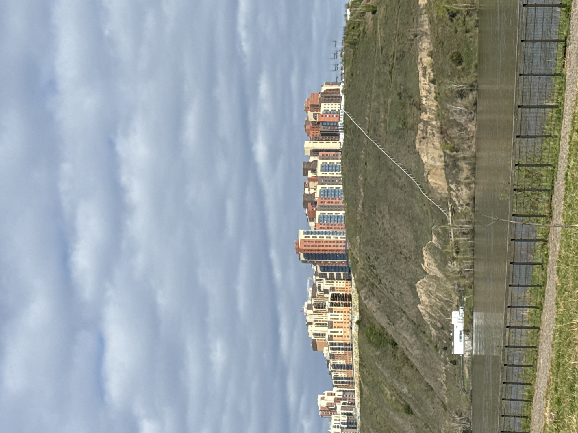 The image depicts a landscape in Krasnoyarsk, Russia, juxtaposing urban development with natural terrain. Along the left, a row of multi-story residential buildings with orange, tan, and blue accents rises against a partly cloudy sky. Adjacent to these buildings, the scene transitions to a natural environment, featuring a steep, grassy hillside marked by patches of exposed earth and vegetation. A service road/walkway is below the hillside. The road is bordered by a metal fence. The landscape is situated against a dynamic sky filled with layers of clouds, indicating a clear and partly sunny day.