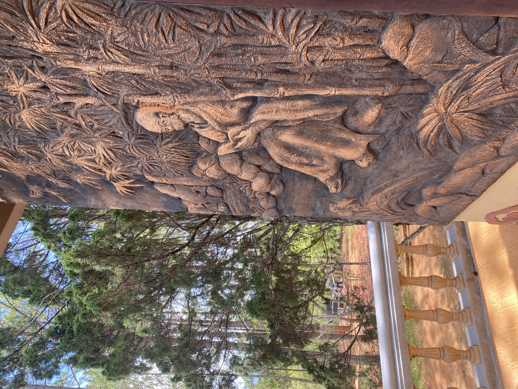 The image shows a stone relief carving depicting a scene of people in a forest, likely a religious or mythological narrative. The carving is highly detailed with figures in robes, trees, and foliage. The carving is located outdoors and appears to be part of a building structure, as evidenced by the adjacent wooden roof and balustrade.
Through the trees, the image shows the city of Da Lat, Vietnam.