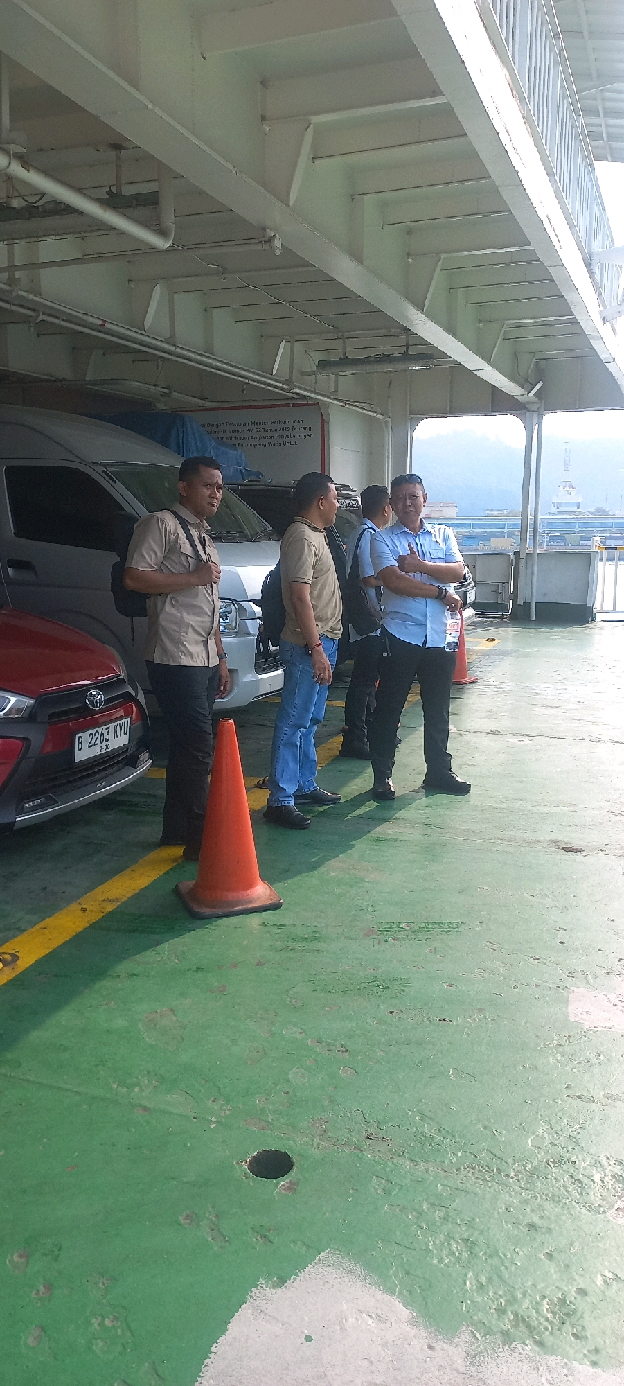 The image depicts a group of four men standing on the deck of a ferryboat in Cilegon, Indonesia. The deck is painted green, with yellow markings and a dark hole visible in the foreground. Two orange traffic cones are placed on the deck. In the background, a white van and a red car are parked, along with part of the ferry's structure, including metal beams and railings. Mountains are visible in the distance. The men appear to be observing something outside the frame. One man on the left wears a tan collared shirt and dark pants, with a backpack. Next to him, a man in a beige shirt and blue jeans stands facing away from the camera. The third man from the left is wearing a light blue collared shirt and dark pants, with a bottle of water in his hand. The fourth man is partially visible on the far right. The scene suggests a moment of pause or waiting on a ferry crossing.