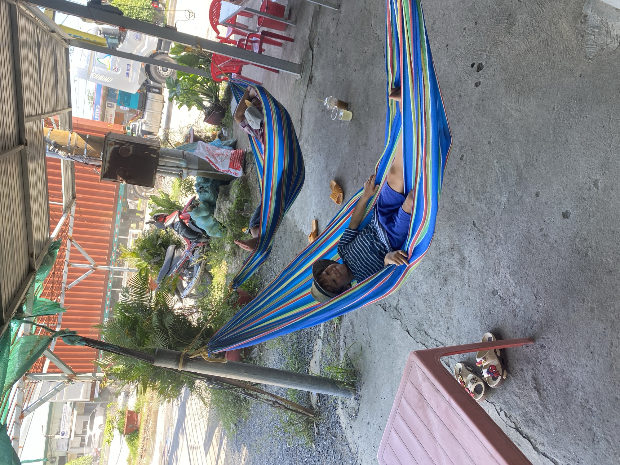 A young boy wearing a striped shirt, blue shorts, and a hat is lying in a colorful striped hammock. His eyes are closed, and he appears to be relaxed or asleep. Another person, whose face is not visible, is lying in a hammock behind him. Both hammocks are suspended between metal poles under a corrugated metal roof supported by a structure with a green mesh covering. In the background, there is a large orange shipping container on a truck, a white truck with a blue logo, and a gas station with a red and white sign that says "PETRO". Various plants and foliage are present around the area. A pink plastic table and a pair of sandals are to the left of the boy's hammock. The scene is on a concrete ground, suggesting an outdoor location, possibly a roadside rest stop or an informal gathering area in Ben Cau, Vietnam.