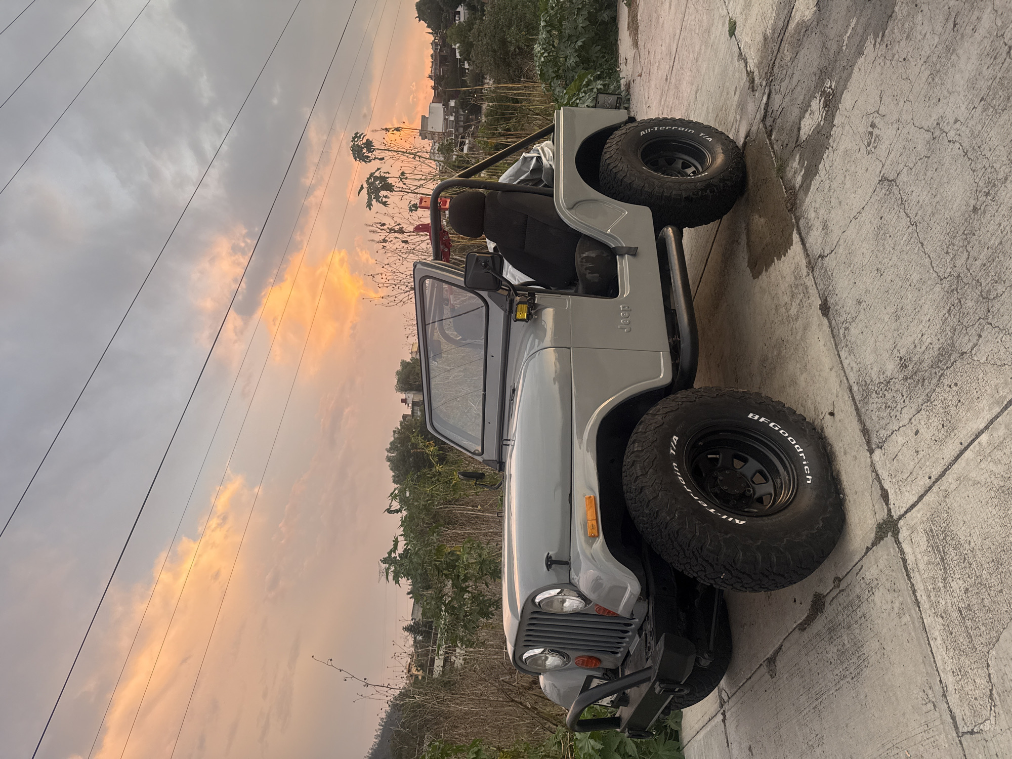 A medium shot depicts a grey Jeep CJ parked on a concrete surface. The vehicle has oversized BF Goodrich All-Terrain T/A tires and a black roll bar. The Jeep's hood is visible, along with the headlights, front bumper, and passenger side mirror. The background features dense vegetation, utility poles with power lines, and a partially obscured view of buildings, suggesting an urban fringe area. The sky is filled with clouds, tinted with hues of orange and grey, indicating either sunrise or sunset. The scene appears to be in Mexico City, Mexico.