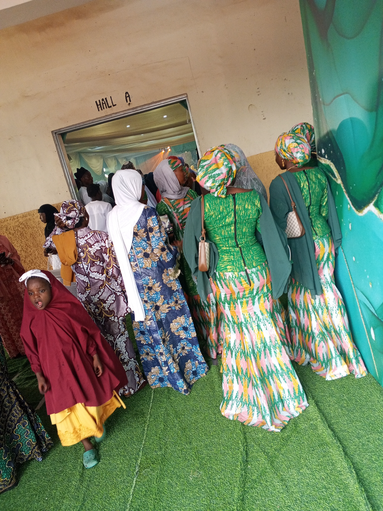 A group of people, predominantly women and a young child, are gathered outdoors near an entrance labeled "HALL A." The individuals are dressed in vibrantly colored, traditional attire, featuring patterned fabrics and headscarves. The ground is covered with artificial green turf. In the background, a large green decorative banner with abstract patterns is visible. The scene appears to be an event or gathering taking place in Abuja, Nigeria, possibly a wedding or cultural celebration. The young child in the foreground is wearing a maroon garment over a yellow skirt and green Crocs, with an open mouth. The other individuals are mostly facing away from the camera, towards the entrance of "HALL A," suggesting they are about to enter or have just exited the hall.