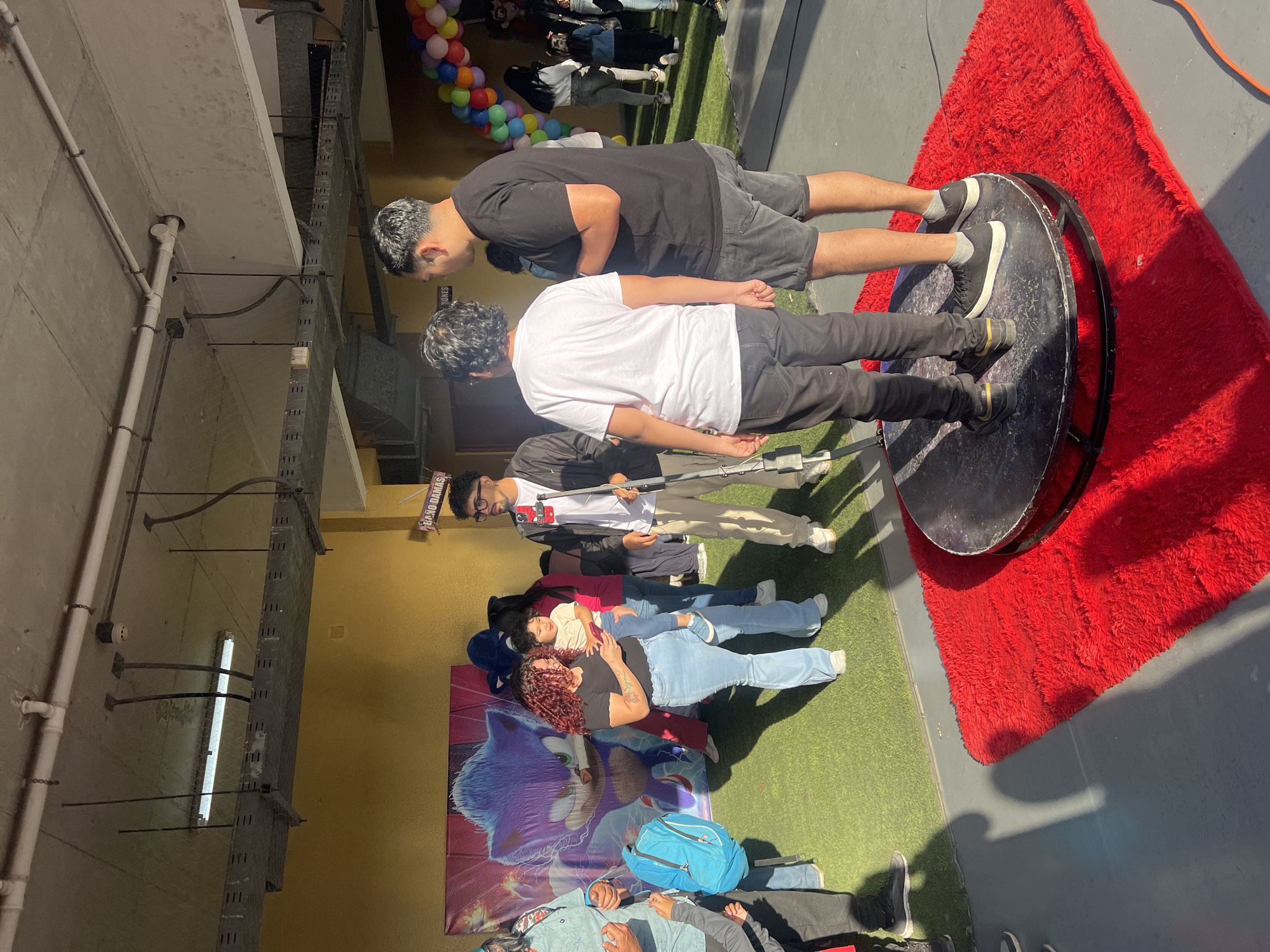 In Arica, Chile, a group of people are gathered for what appears to be a birthday party. In the background, a banner featuring the character Sonic the Hedgehog suggests a themed event. A circular platform covered with a red fluffy rug is set up, with two young boys standing on it, likely for a photo opportunity. Several adults and children are present, with some observing the scene and others participating. One woman is holding a baby, while another individual appears to be operating a camera or recording device mounted on a tripod. The environment seems to be an indoor space with an artificial grass floor, possibly a community hall or event space, with a sunny outdoor area visible to the right. Balloons in various colors are also present, adding to the festive atmosphere.