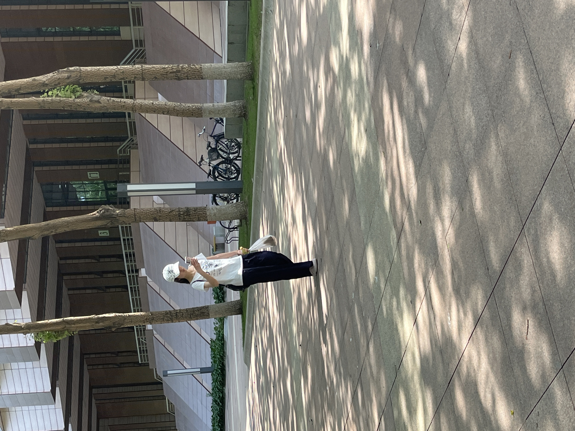 A young woman is walking on a paved path in front of a modern building with tiered walkways. She is wearing a white t-shirt, dark pants, a white baseball cap, and sneakers. She is holding a mobile phone in her right hand and a plastic bag with something inside in her left hand. Sunlight filters through the trees, casting shadows on the ground. Two bicycles are parked behind her near the building's entrance. The building appears to be made of concrete and glass, with light-colored stone accents. The scene is likely a campus or public area in Changchun City, China.