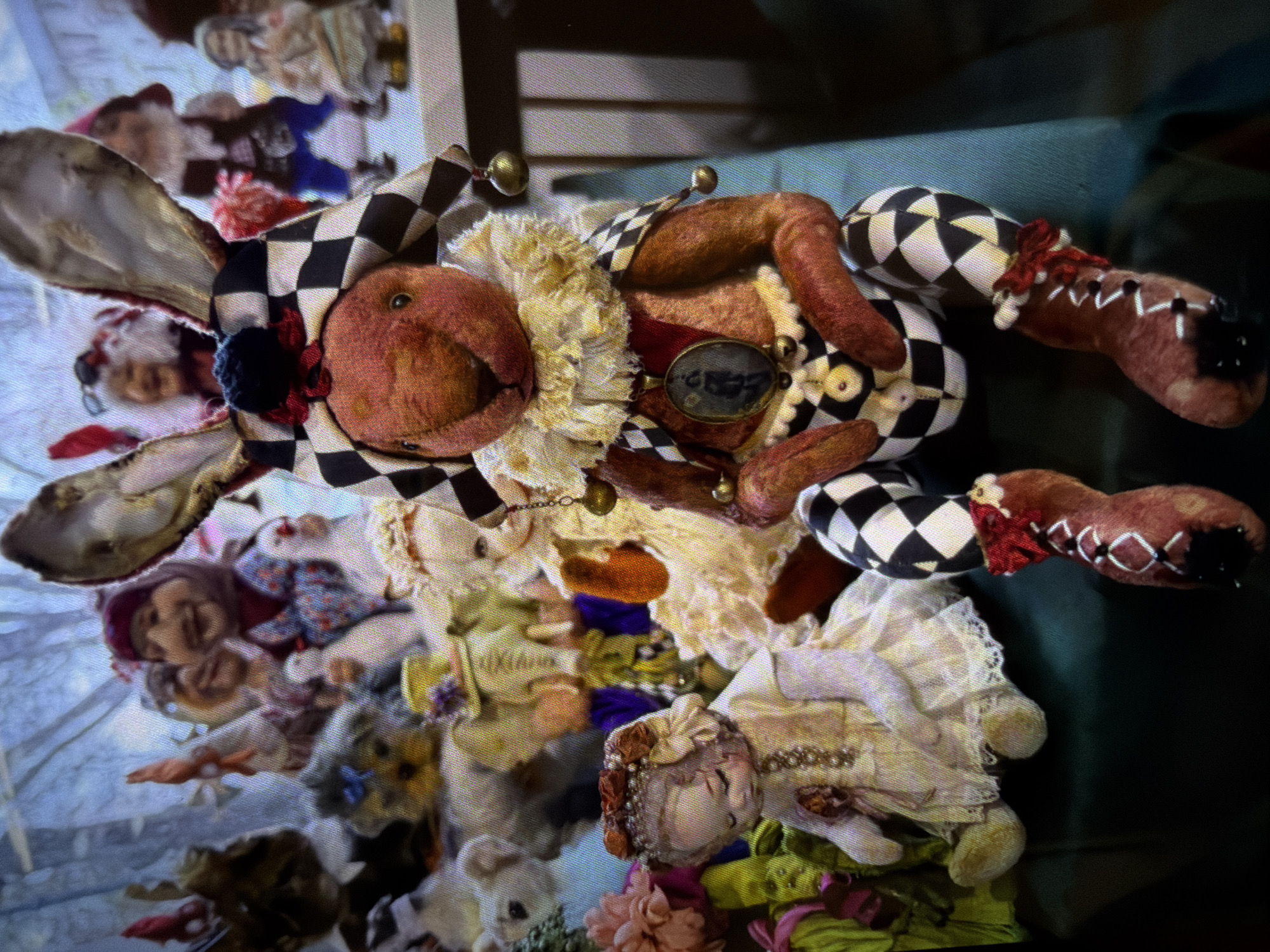 The image depicts a collection of handmade dolls, seemingly arranged for display or storage. Prominently featured is a jester-like doll with a rabbit's head adorned in a black and white checkered pattern hat, complete with dangling bells. This doll is posed atop a pile of other dolls, some with porcelain faces and intricate clothing. The background appears to be a cluttered area, possibly a craft room or storage space. The location in the city of Moscow, Russia cannot be determined from this image.