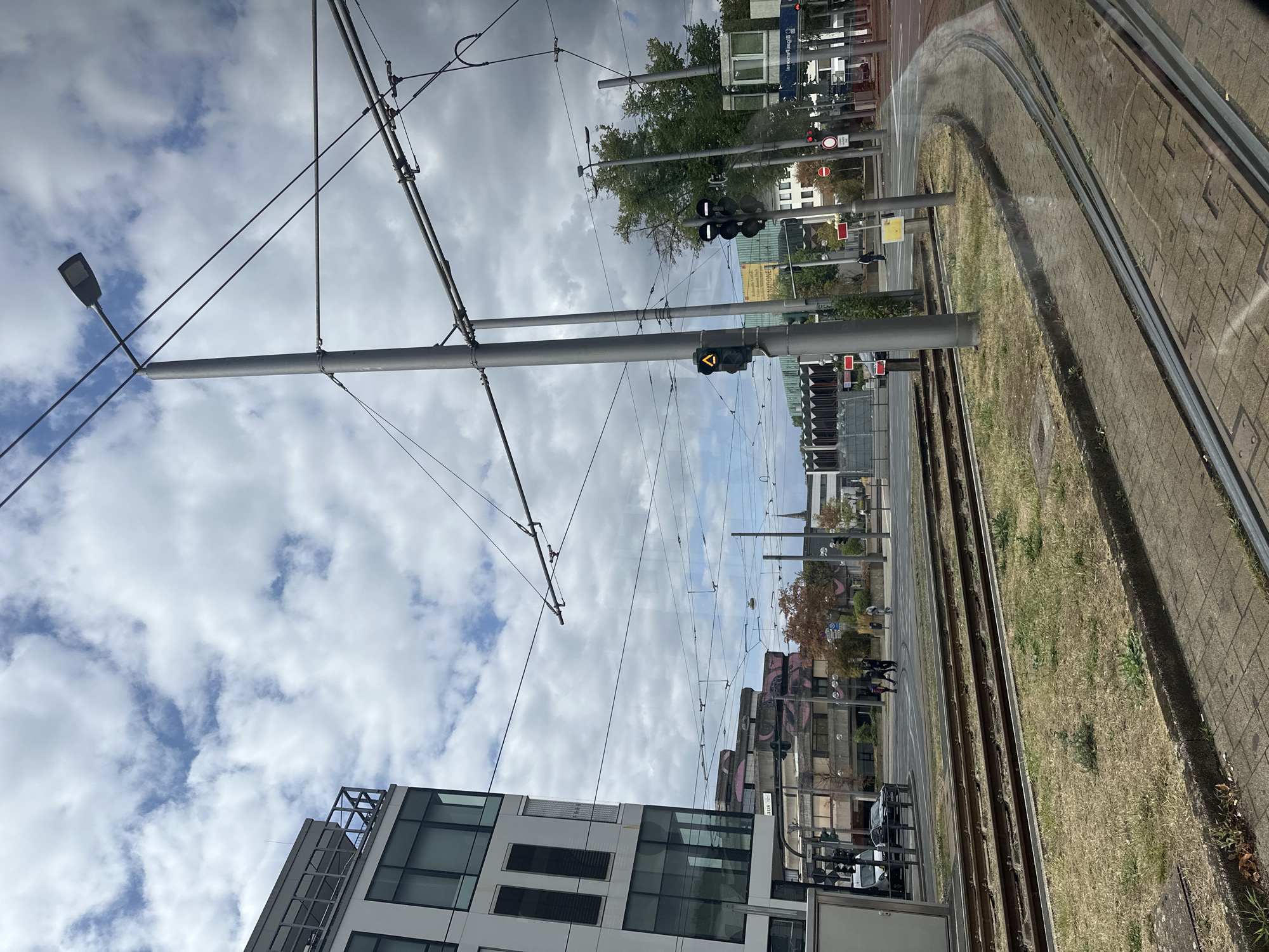 The image depicts a view from a tram in Krefeld, Germany, looking towards a street intersection and surrounding buildings. The foreground features tram tracks, some overgrown with grass, and a paved sidewalk.
In the mid-ground, there is a street with a few vehicles and pedestrians crossing. Traffic lights are visible, with one displaying a yellow arrow. Several tall metal poles with overhead wires for the tram are prominent throughout the scene.
To the left, a modern multi-story building with large windows dominates the view. Further in the background, other buildings of various architectural styles are visible, including one with a distinctive pink mural. Trees with autumn foliage are present in the distance. The sky is partly cloudy.