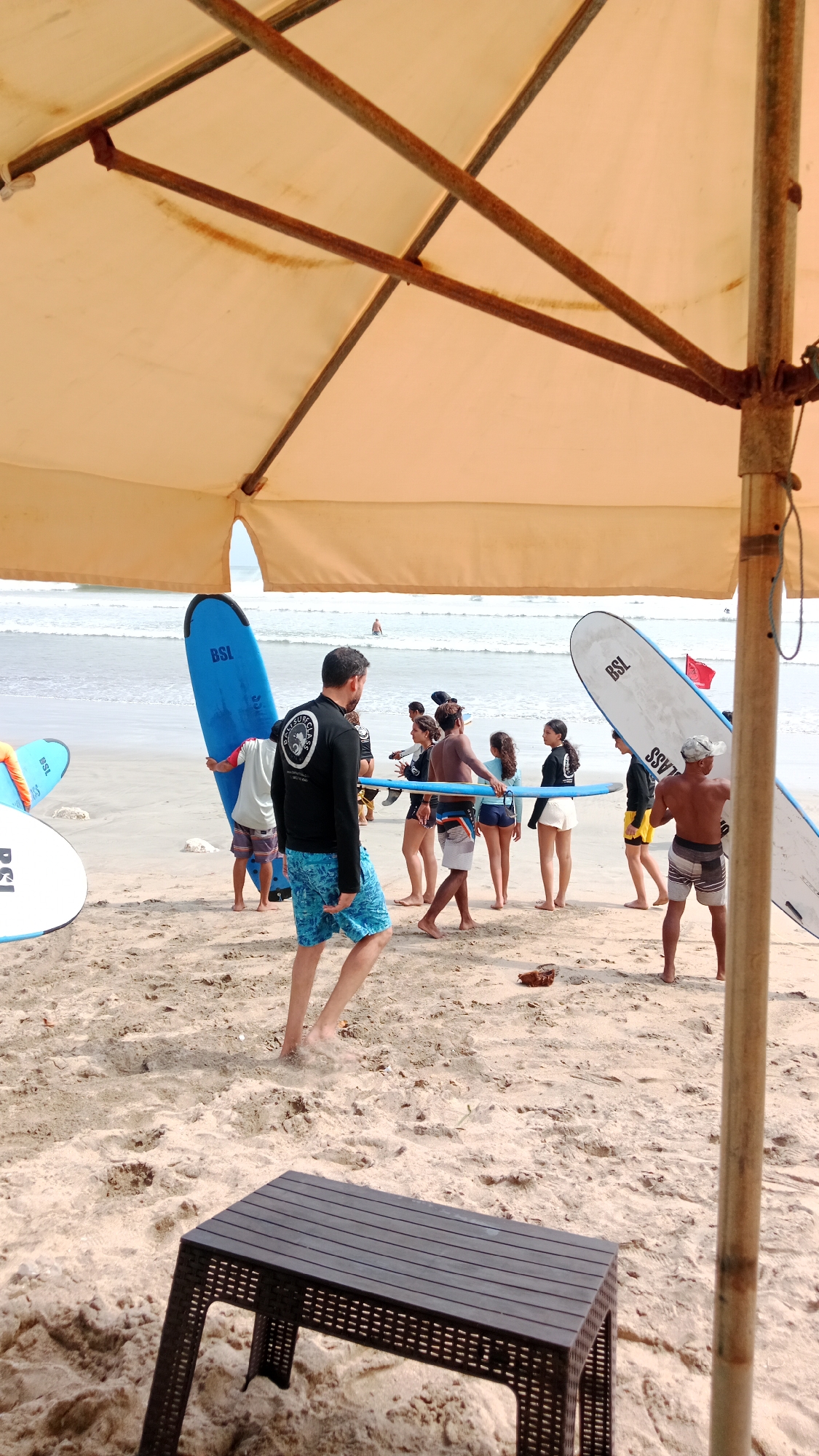 A group of people are gathered on a sandy beach near the ocean, with some holding surfboards. The scene is partially covered by a large, beige umbrella with visible metal supports. In the foreground, a dark brown, slatted plastic table sits on the sand. Several individuals, including men and women, are dressed in swimwear and rash guards, some of whom appear to be participating in a surfing lesson. Two blue surfboards are leaning upright on the sand, and another white and blue surfboard is held by one of the individuals. In the background, gentle waves are visible in the ocean, and a single person is in the water. The lighting suggests it is daytime.