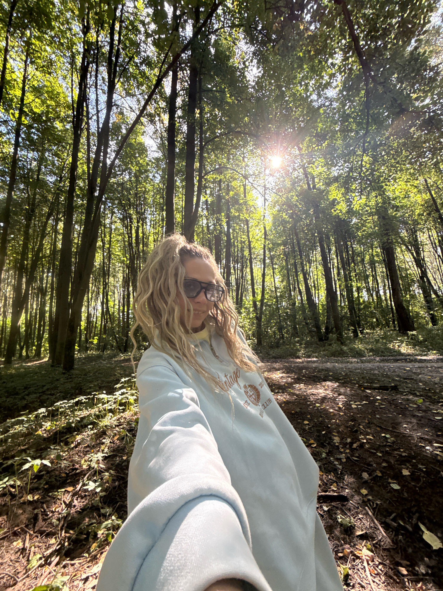 In the city of Dumino, Russia, a young woman is captured in a selfie taken within a sun-dappled forest. The woman, with blonde, curly hair pulled into a high ponytail, wears dark sunglasses and a light blue sweatshirt with a graphic design on the chest. She is positioned in the foreground, her body angled towards the camera, while the background showcases a dense collection of tall trees. Sunlight streams through the canopy, creating bright patches on the forest floor, which is covered with leaves and small plants. The overall impression is one of a casual outdoor excursion in a natural setting.