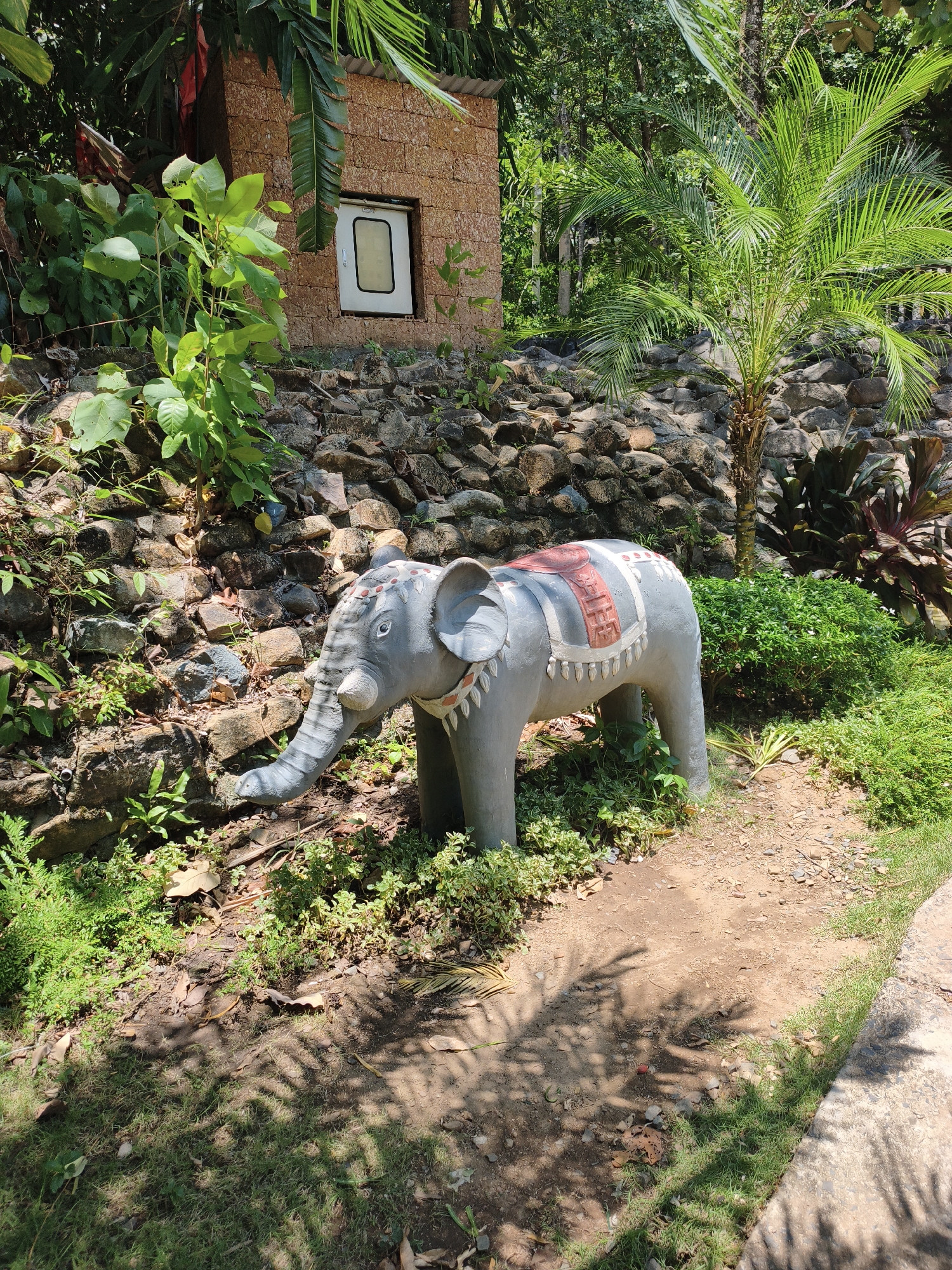 In Tuy Hoa, Vietnam, an elephant statue, adorned with a decorative blanket, stands on a dirt path beside a stone wall and lush green foliage. The statue, rendered in a neutral gray, features intricate patterns on its head and blanket. Behind it, a small, rustic building constructed from brown stone-like material is partially visible, featuring a white-framed window. The environment is characterized by dense vegetation, including palm trees and various shrubs, under a bright, sunny sky that casts dappled shadows on the ground.