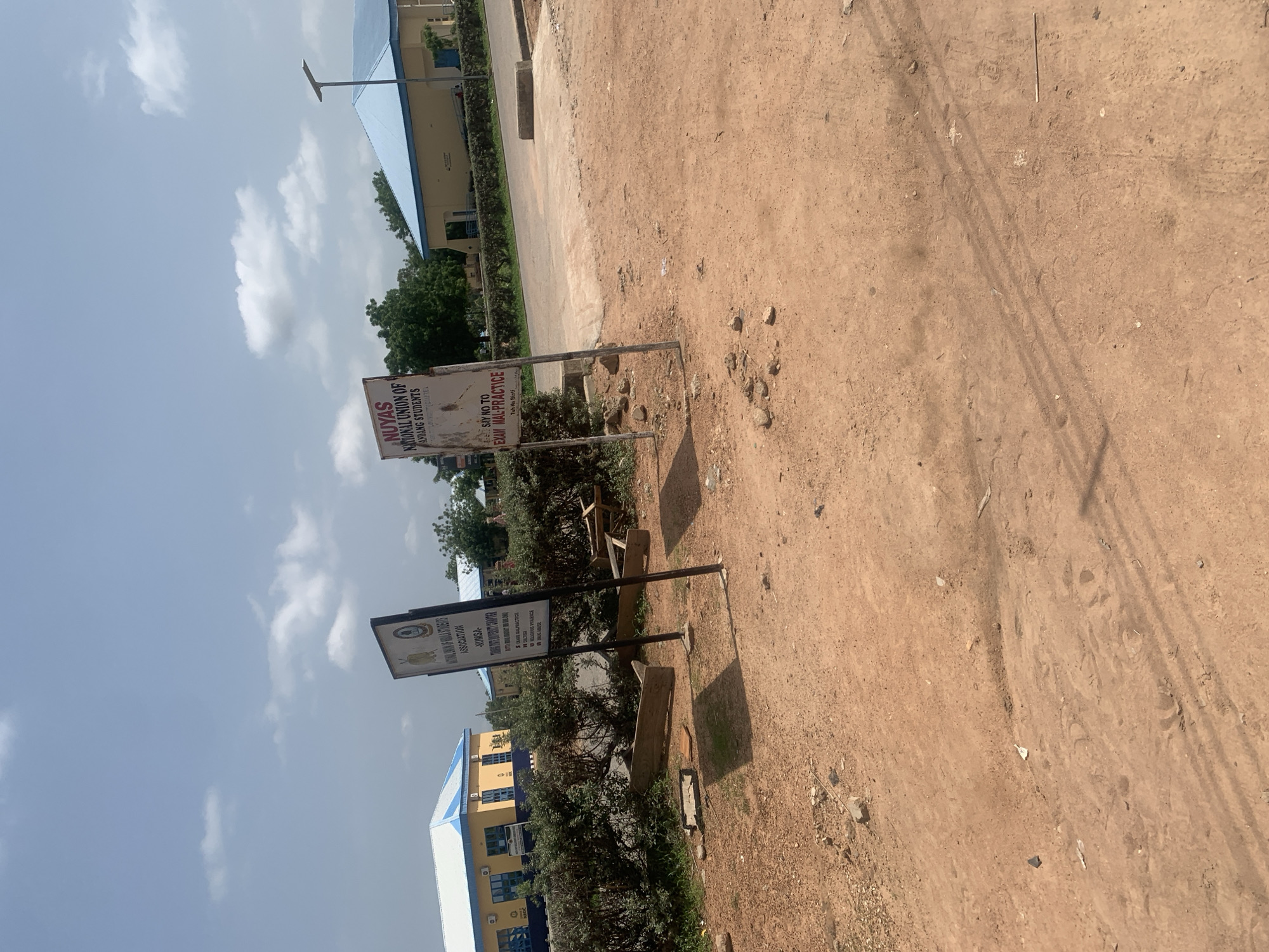 The image depicts an outdoor scene with two signs on posts in the foreground, located on a dirt path. The sign on the left has a white background with text and a logo, appearing to be a notice board or informational sign. The sign on the right is also a notice board with text, and it's partially obscured by the other sign and shadows. Behind the signs, there is a row of green bushes and trees. In the background, there are two modern-looking buildings with blue roofs, suggesting an institutional or educational setting. A street lamp is visible on the right side of the frame. The ground in the foreground is unpaved and covered in reddish-brown dirt with scattered rocks and debris. The sky is partly cloudy. This scene is located in Jalingo, Nigeria.