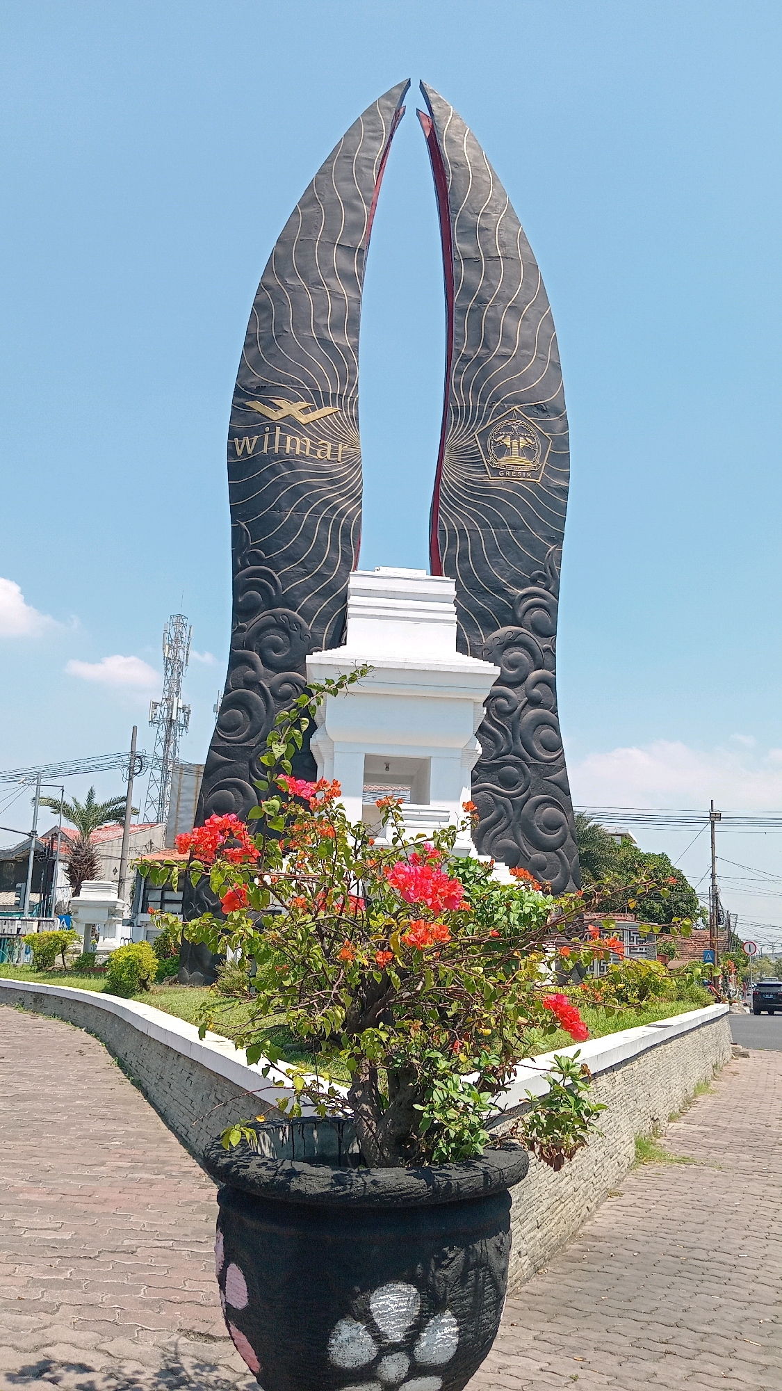 This is a vertical, eye-level, outdoor, daytime, full shot of a monument in Gapurosukolilo, Indonesia. The monument is a large, black, abstract structure with a red interior. It is composed of two large, curved elements that meet at the top, resembling open petals or wings. The surfaces of these elements are decorated with intricate, swirling gold lines. To the left of the monument, the word "wilmar" is visible in gold lettering. To the right, a circular emblem with gold lettering that reads "GRESIC" is present.
In front of the monument, a white, tiered structure stands, appearing to be a smaller pedestal or shrine. In the foreground, a large, black planter contains a vibrant bush with numerous bright red and orange flowers. The planter has a decorative pattern of light-colored circles.
The monument is situated in a paved area with a curved walkway made of interlocking bricks. A low, stone retaining wall runs alongside the walkway. Beyond the monument and planter, the environment includes a clear blue sky with scattered white clouds. In the background, a tall cellular tower is visible, along with electrical poles and wires, some buildings, and trees. A black car is driving away on the right side of the road. The overall scene suggests a public space, possibly a roundabout or a park entrance, within an urban or semi-urban setting.