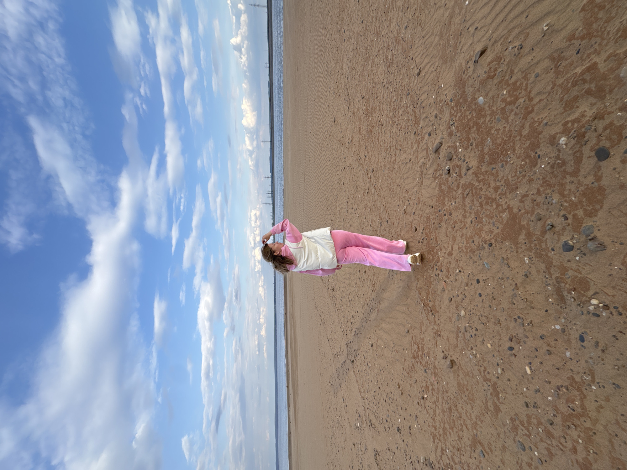 A woman lies on a beach. She is wearing pink sweatpants and a white vest. Her arms are raised above her head. The beach is sandy, and there are small stones scattered throughout. Beyond the beach is the water, and beyond the water is a narrow stretch of land. There are pylons running along the narrow stretch of land. The sky is blue, and there are white clouds. The image was taken on a beach in the city of Chaikovsky, Russia.