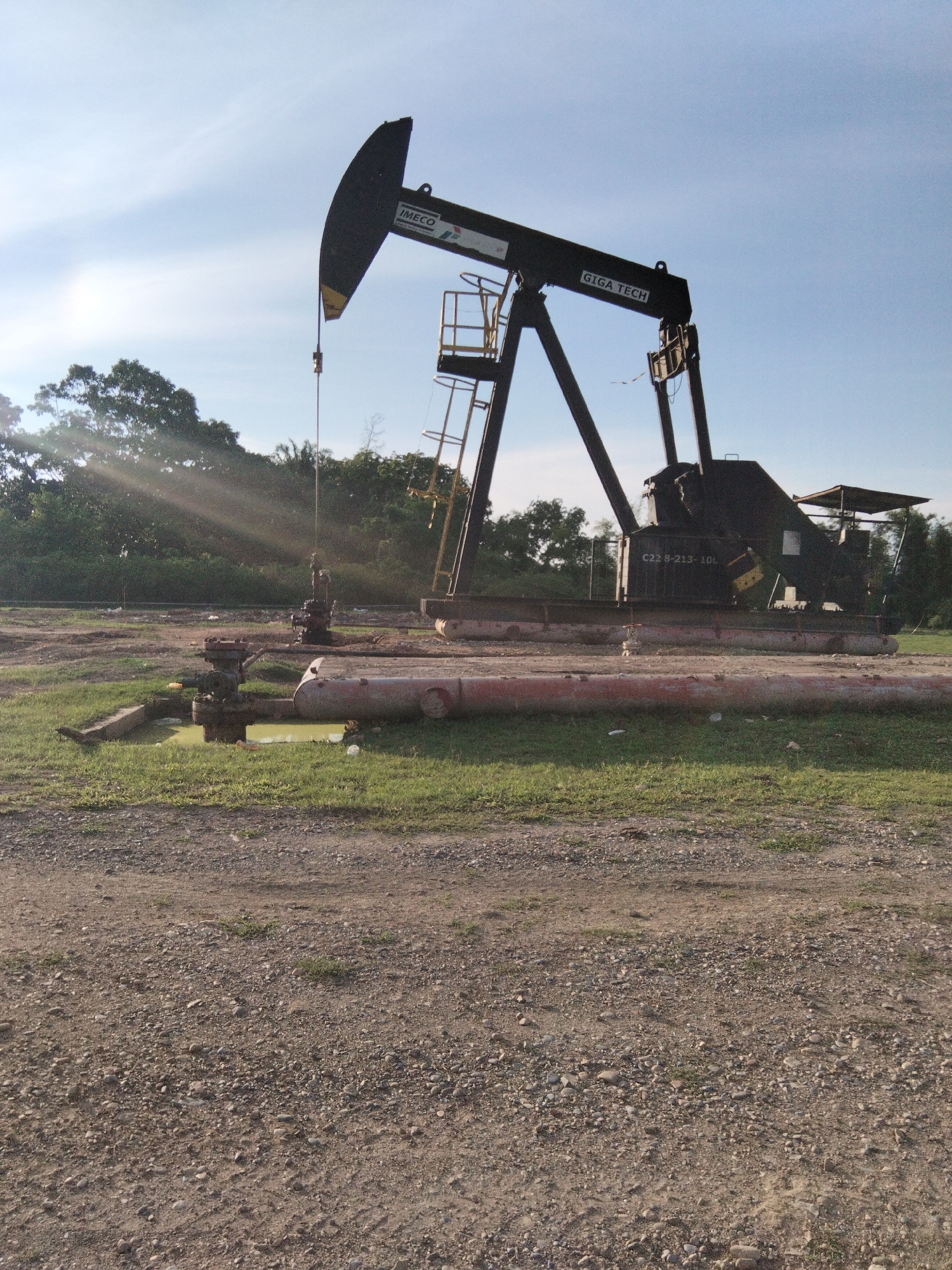 The image displays an oil pumpjack in an outdoor setting, likely an oil field. The pumpjack is predominantly black and features the logos "IMECO" and "GIGA TECH" on its beam. A ladder is attached to the structure. In the foreground, there are several industrial pipes, some of which appear to be resting on the ground and are red and metallic in color. A pool of yellowish liquid, possibly oil or wastewater, is present near some of the pipes. The ground is a mixture of dirt, gravel, and sparse grass. In the background, lush green trees form a dense line, and the sky is a clear blue with some wispy clouds and visible sun rays. There are no people or animals depicted in the image. The scene suggests an operational or recently operated oil extraction site. The location is identified as Sukaramai Dua, Indonesia.