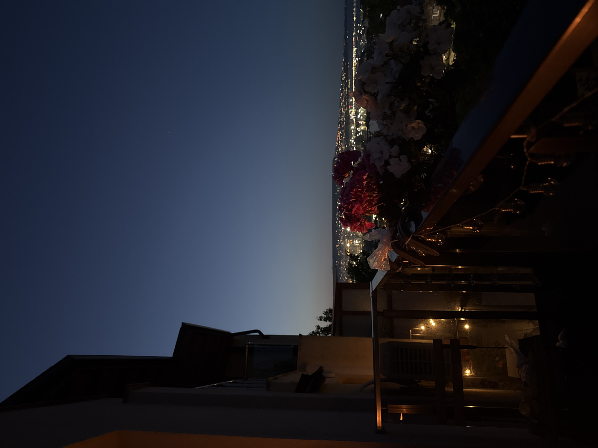The image depicts a balcony at dusk, overlooking a city skyline dotted with lights. On the balcony, there are flowers, some of which are pink and white, and string lights. To the left of the balcony, part of a building is visible, with warm light emanating from a window. The sky is a deep blue, transitioning to a lighter hue near the horizon. The city lights suggest an urban environment, possibly with a body of water in the distance. There are no people visible in the image.