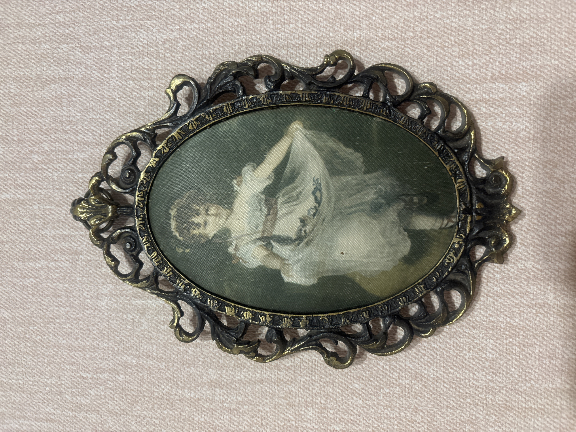 A framed painting of a young girl in a white dress is displayed on a pink textured wall. The frame is ornate and made of dark metal with gold accents. The painting depicts a girl with dark hair, wearing a full, flowing white dress with a sash and delicate detailing. She is holding up the skirt of her dress with her left hand, and her right arm is bent, with her hand near her waist. The background of the painting is a muted green. The overall aesthetic suggests a vintage or antique piece. There is no information available about the specific location of this image within Yanino-1, Russia.