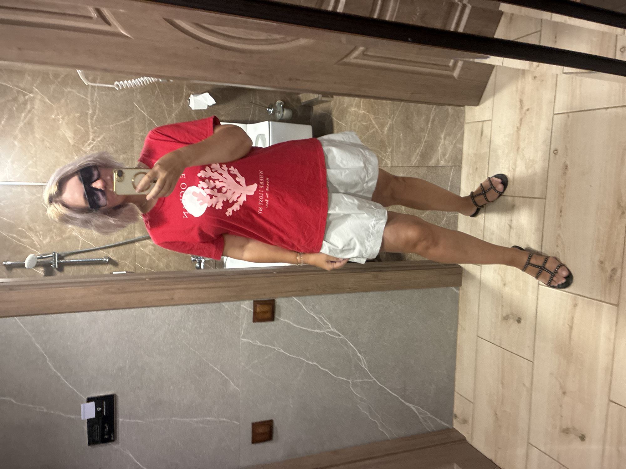 A woman with blonde hair and sunglasses stands in a tiled bathroom, taking a mirror selfie. She is wearing a red t-shirt with a coral graphic and white shorts. She has on black sandals. The bathroom has marble-patterned walls and light wood-colored floor tiles. A shower head is visible on the wall. The woman appears to be in a hotel or similar accommodation in Estosadok, Russia.