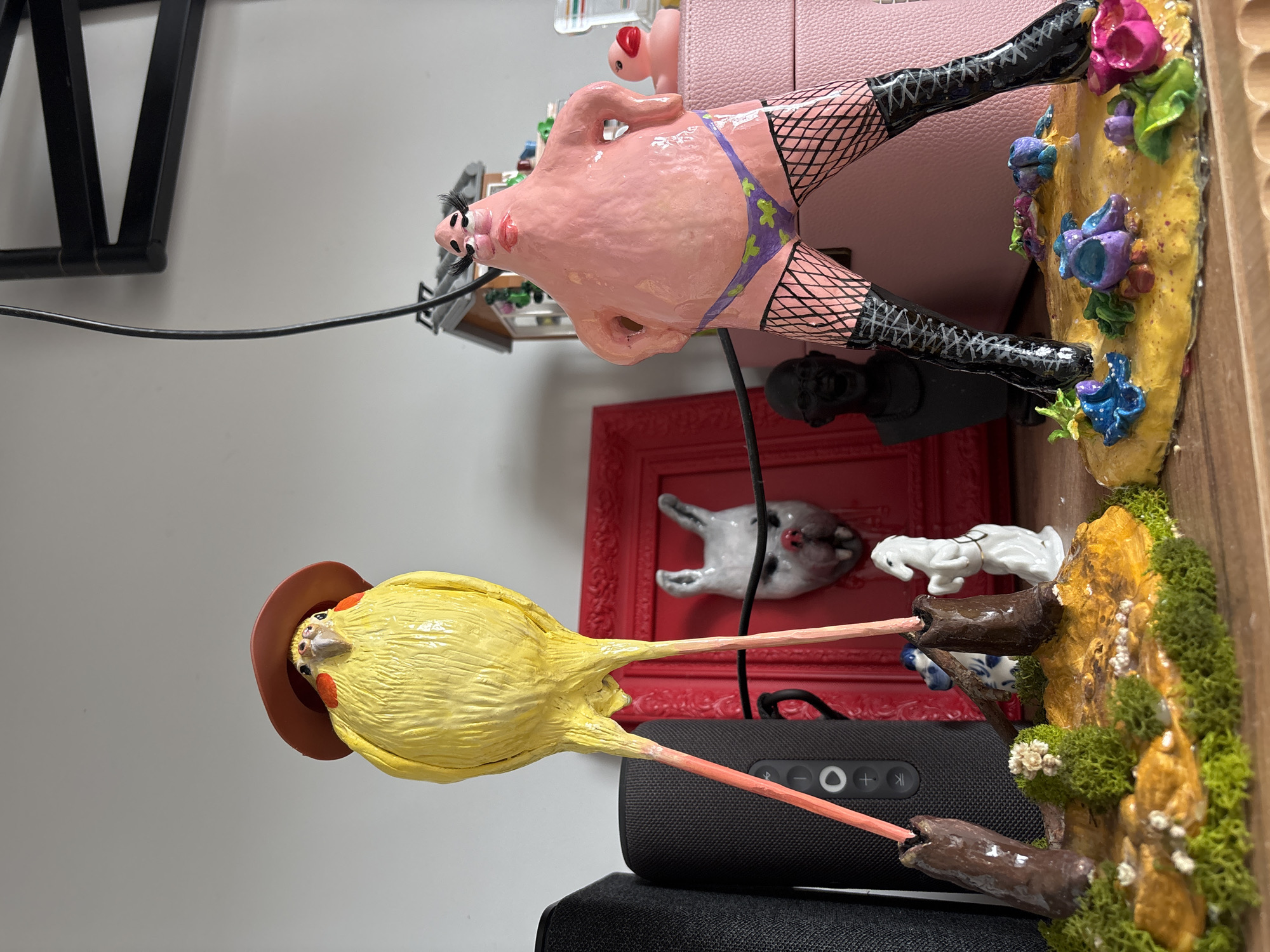 The image shows a collection of eclectic figurines on a shelf or desk. In the foreground, there is a yellow bird figurine with a cowboy hat. Behind it is a dark gray speaker. Above these items, there is a red picture frame, small cat and dog figurines, and a pig figurine in fishnet stockings and a bikini. The pig figurine is positioned next to a pink purse or bag. The background is a white wall with a black metal frame structure visible in the upper left. There is no information in the image to ascertain that it is in Krasnoyarsk, Russia.