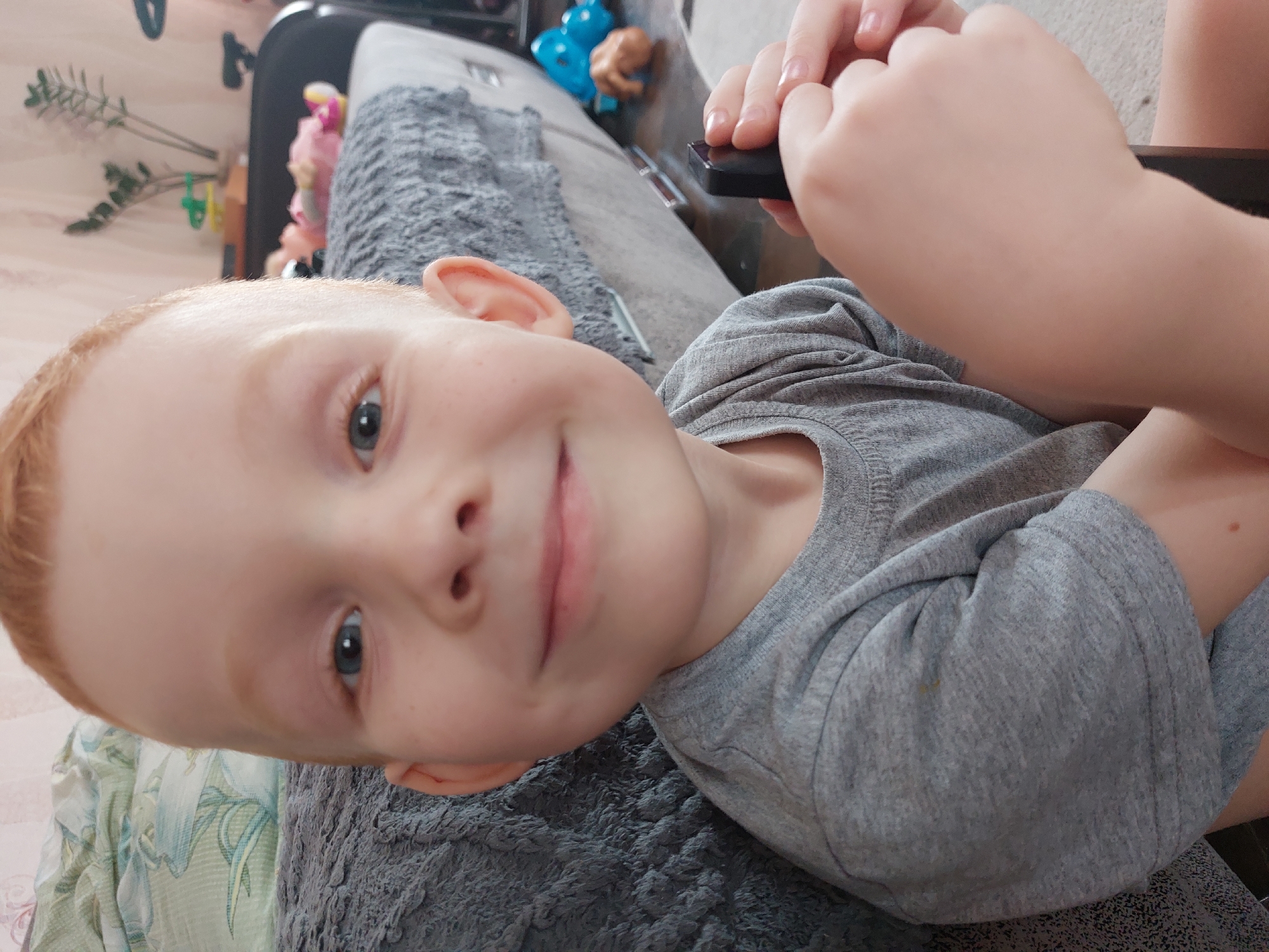 A fair-skinned child with reddish hair is lying down. The child is wearing a gray shirt. In the upper right, a person's hands are visible, holding a black object. The child is smiling. A gray textured surface is beneath the child. Behind the child's head are indiscernible objects on a surface. The location is unknown.