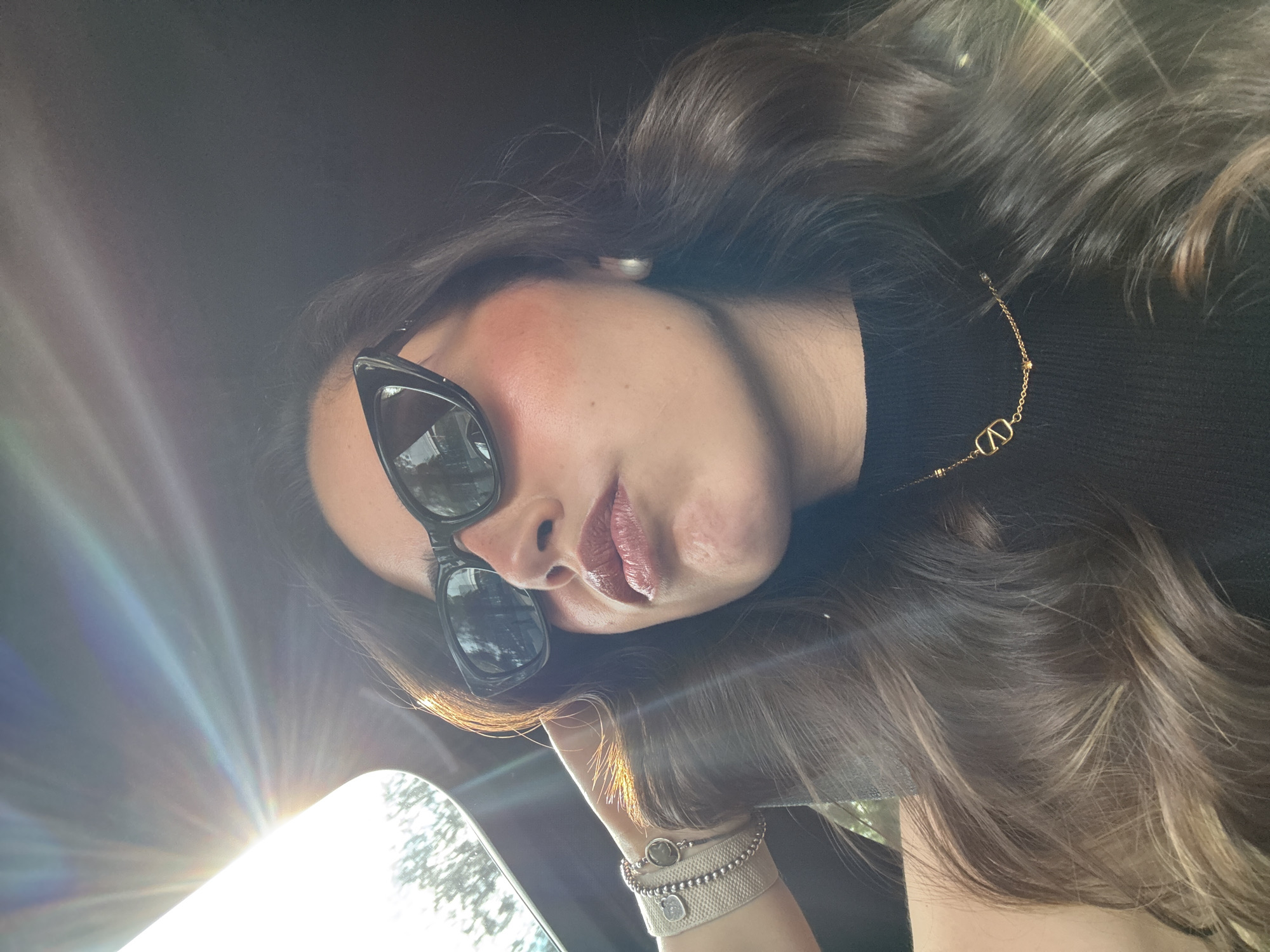 A low-angle, selfie-style shot depicts a woman with dark hair and sunglasses, possibly taken inside a car. The sun is shining brightly, causing a glare near the rearview mirror in the lower left corner. The woman wears a black top and a gold necklace. Bracelets adorn her wrist, which rests near the mirror. The location in the city of San Bernardino Tlaxcalancingo, Mexico, cannot be determined from the visual information provided.