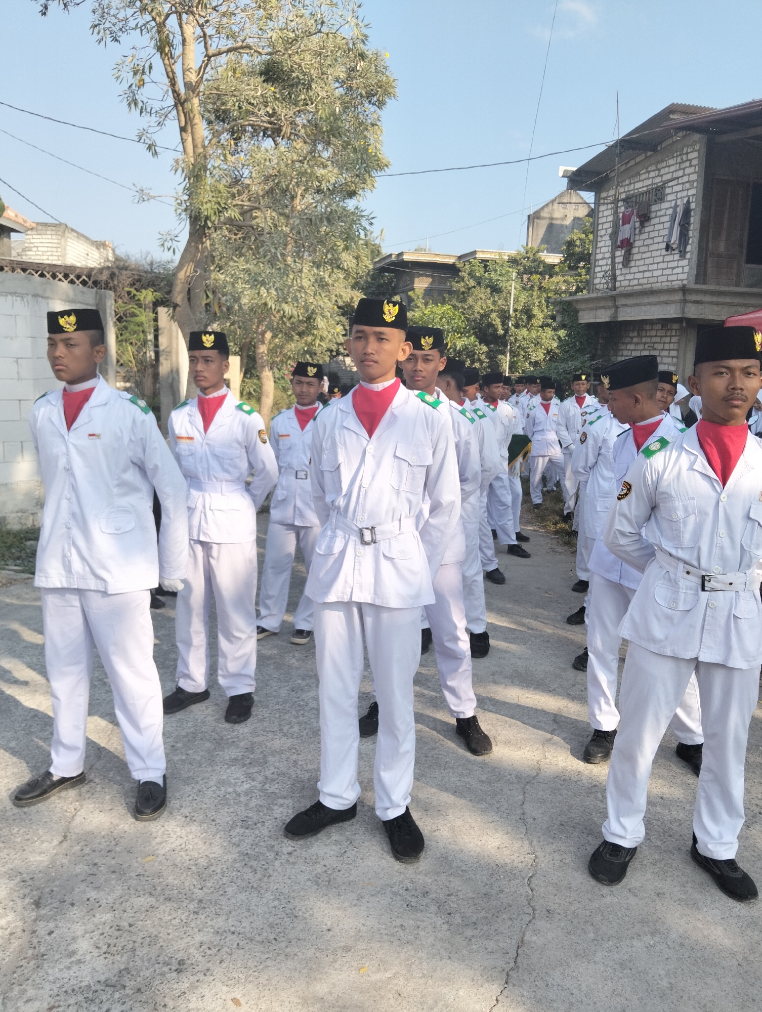 A group of young males, likely students, stand in formation outdoors in what appears to be an urban residential area in Paciran, Indonesia. They are all wearing identical white uniforms consisting of long-sleeved shirts with chest pockets and epaulets, white trousers, black hats with golden emblems, and red neckerchiefs. Most of the individuals are also wearing black shoes. The background features a street or paved area, a mature tree with sparse foliage, and the side of a two-story building constructed of white brick. A fence is visible to the left of the building. The sky is clear and blue. The scene appears to be captured during daylight hours.