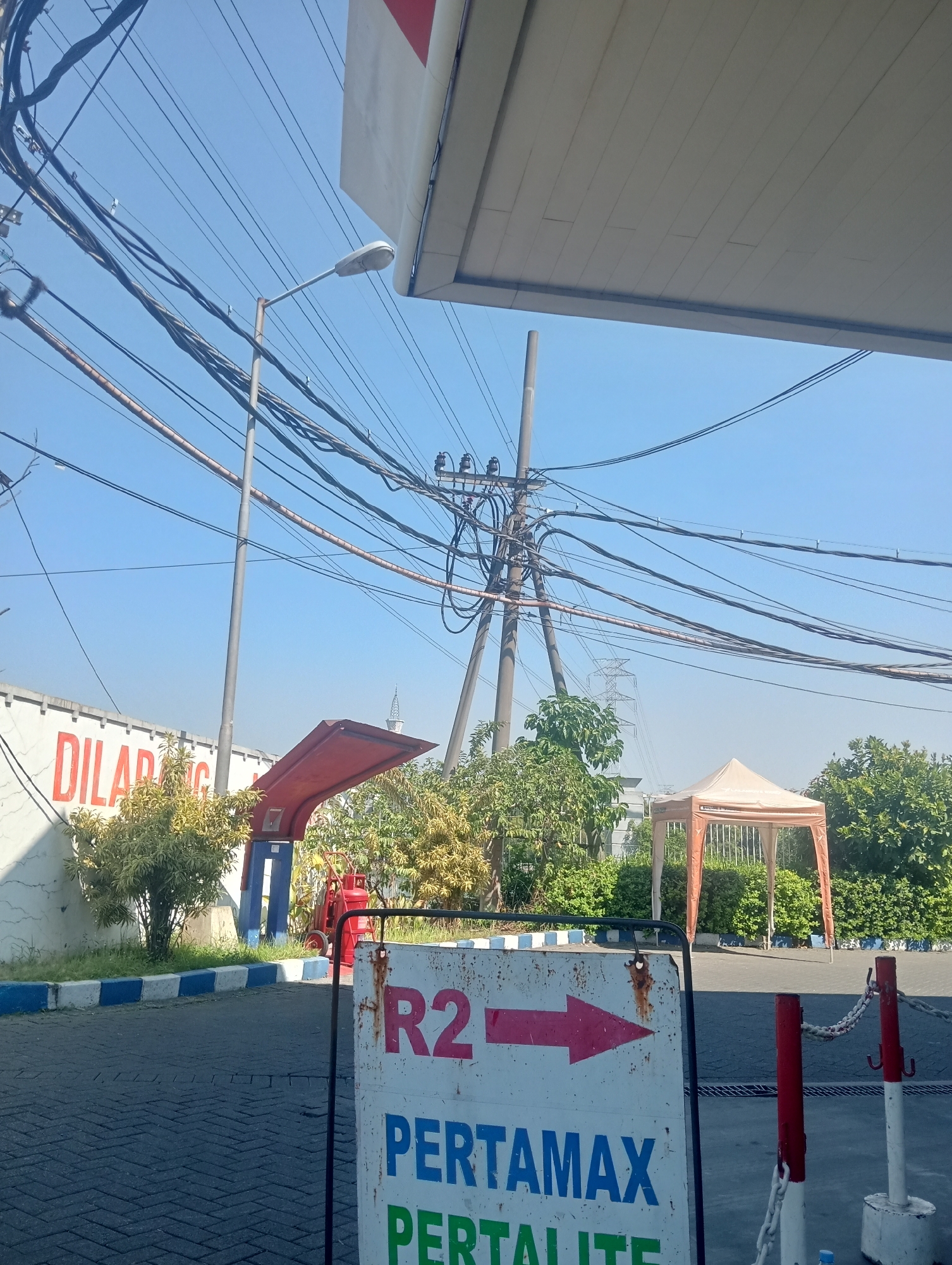The image captures an outdoor scene, likely at a gas station in Surabaya, Indonesia, under a clear blue sky. A large, white canopy with a red stripe extends from the top of the frame, suggesting it's part of the gas station's structure. The foreground features a grey paved surface, possibly a road or parking area, with a sign on the right indicating "R2" and an arrow pointing right, followed by text that appears to read "PERTAMAX PERTALITE". This sign is weathered and rusted.
To the left, a white wall with large red lettering that reads "DILARANG" (meaning "forbidden" in Indonesian) is visible. Behind this wall, there are lush green trees and bushes. A red fire extinguisher on wheels is also present. Further in the background, a small, orange tent or canopy stands, and beyond that, dense green foliage and a tall electrical pylon with numerous wires are prominent against the sky. A street light is also visible amidst the tangle of electrical wires. The overall impression is a typical roadside scene in an Indonesian city, likely a service area.
