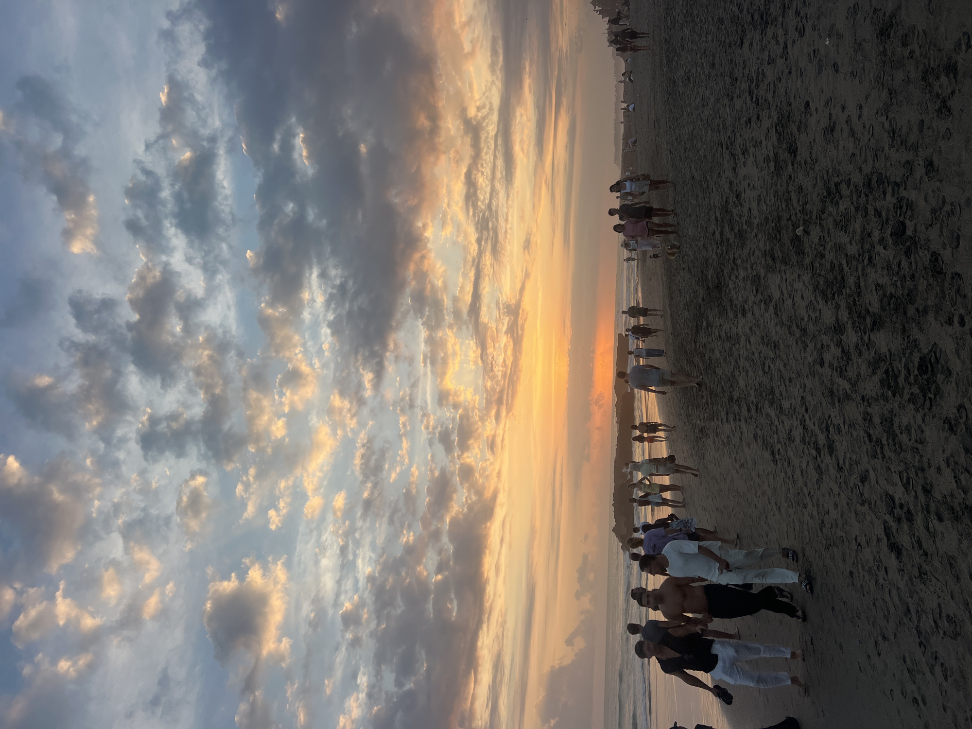 This is a wide shot of a beach at sunset in North Kuta, Indonesia. The sky is filled with dramatic clouds tinged with orange and yellow from the setting sun, with patches of blue sky visible. The ocean is calm with small waves. A group of people are walking along the sandy beach, some in the foreground and others further in the distance. The people are casually dressed in beach attire. The sand is dark and appears wet in some areas, with footprints visible. In the distance, there are some structures that could be buildings or other beach amenities. The overall scene evokes a sense of a popular beach destination during a beautiful sunset.