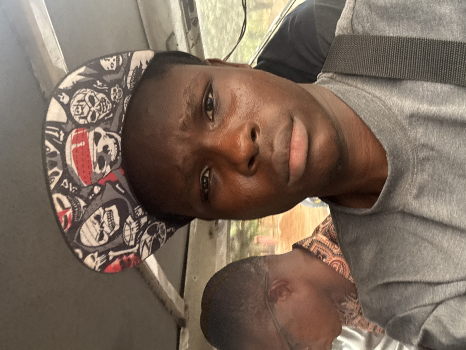 A dark-skinned man wearing a grey t-shirt and a skull-patterned baseball cap is angled to the right. A black strap goes over his shoulder. His facial expression is neutral. Below him is another person with short hair wearing glasses and a patterned shirt. It is unclear who they are. Outside a window, there is green foliage.