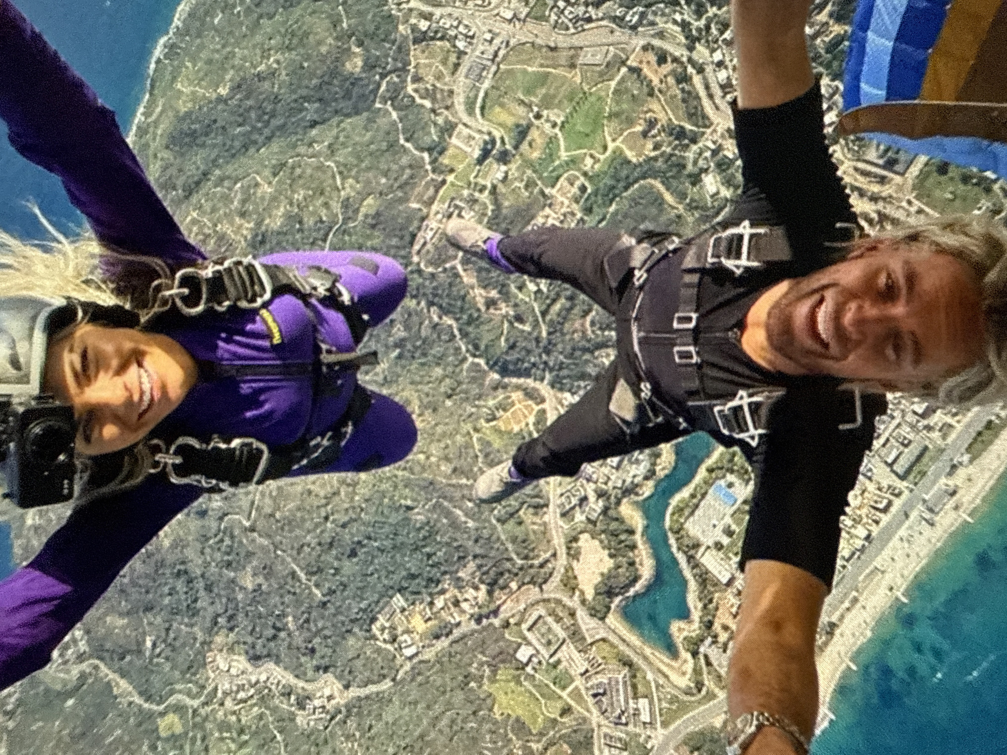Two individuals are captured in a mid-air skydiving scene. The person on the left is wearing a purple jumpsuit and a helmet with a camera attached, while the person on the right is wearing black clothing and has a watch on their wrist. Both individuals are smiling and facing the camera. The background reveals an aerial view of a coastal region with a mix of vegetation, buildings, and bodies of water. The shoreline is marked by a beach and urban development. A partial view of a blue and white parachute is visible in the upper right corner. The location is Unknown, Unknown.