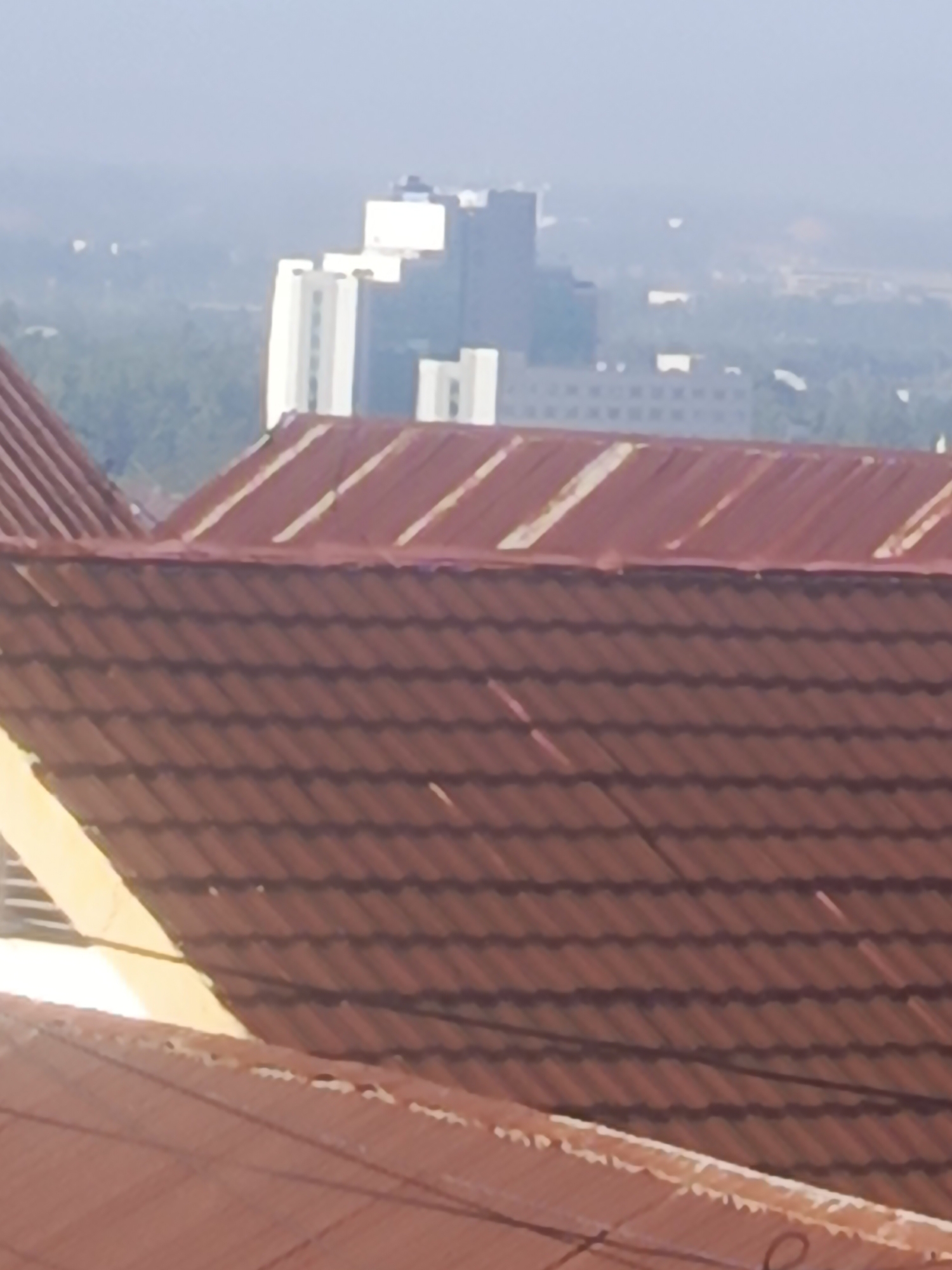 The image captures a hazy, distant cityscape in Kendari, Indonesia. In the foreground, two tiled roofs dominate the view, with the closer one featuring reddish-brown tiles and a yellow trim on the left side. The roof further back, appears to be made of corrugated metal painted reddish-brown with lighter colored ridges. Beyond the roofs, a cluster of buildings is visible in the mid-ground, including several white, modern structures and a taller, multi-story building with a stepped facade. The background is obscured by atmospheric haze, with hints of greenery and possibly more distant structures. The overall impression is of a sunny day with poor visibility.